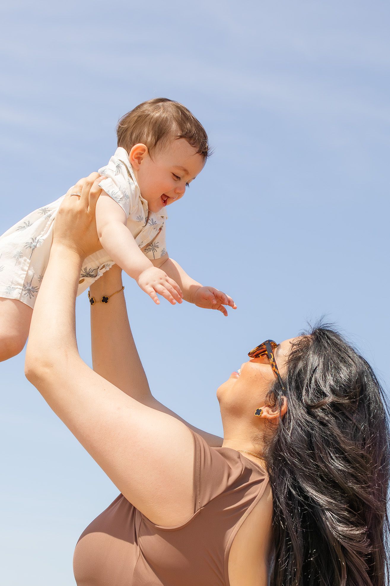 A joyful mother lifts her smiling baby high against a clear blue sky, sharing a playful outdoor moment.