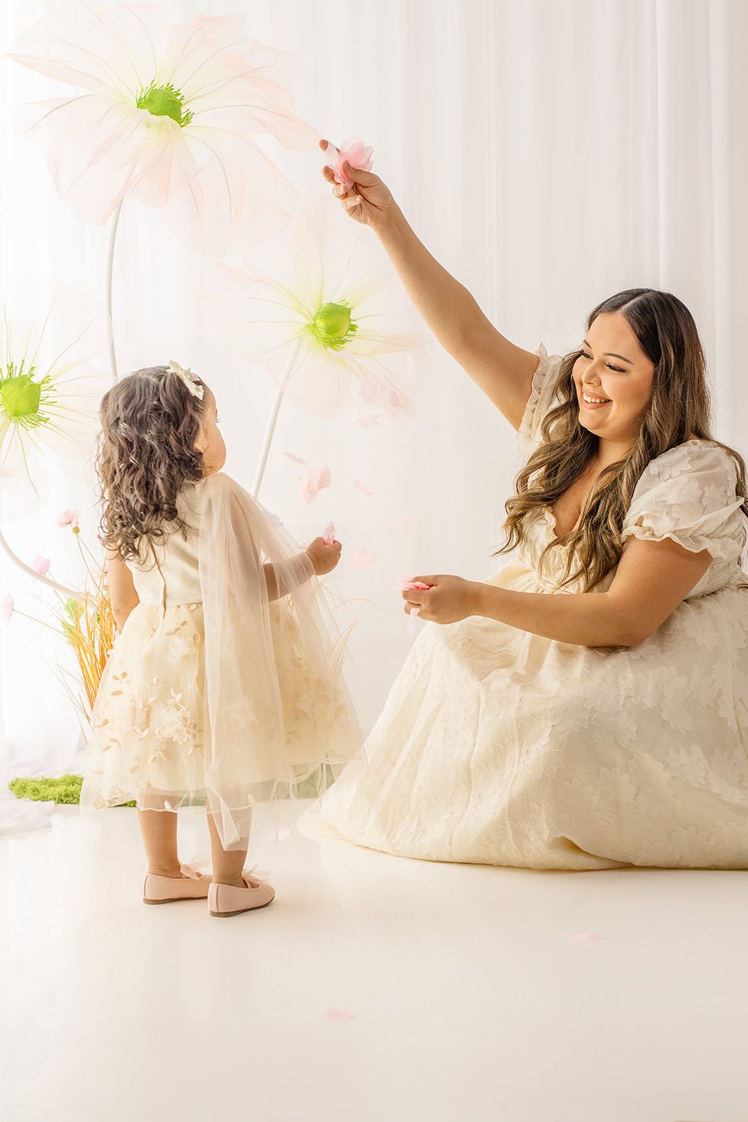 Mother and daughter in white dresses playing with oversized flowers in a bright, whimsical studio setting.