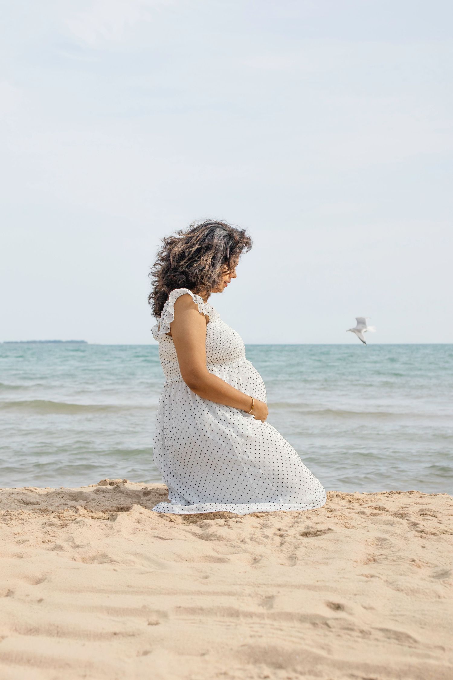 A pregnant person in a white polka dot dress sits peacefully on a sandy beach by the ocean.