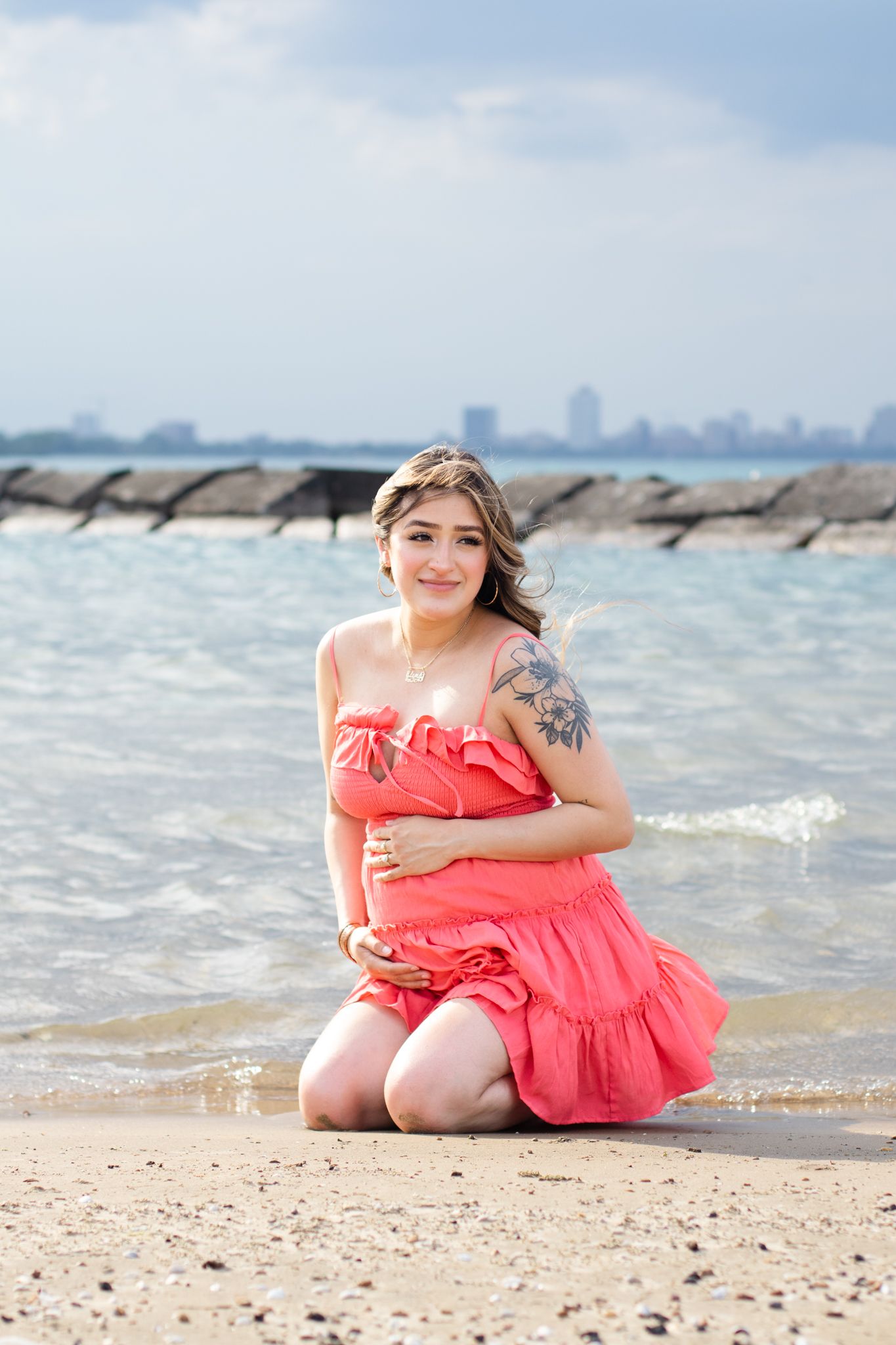 Woman in coral dress poses on sandy beach with city skyline visible in background.