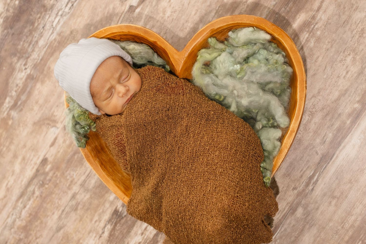 Sleeping newborn wrapped in brown blanket lying in heart-shaped basket with mint green padding on wooden floor.