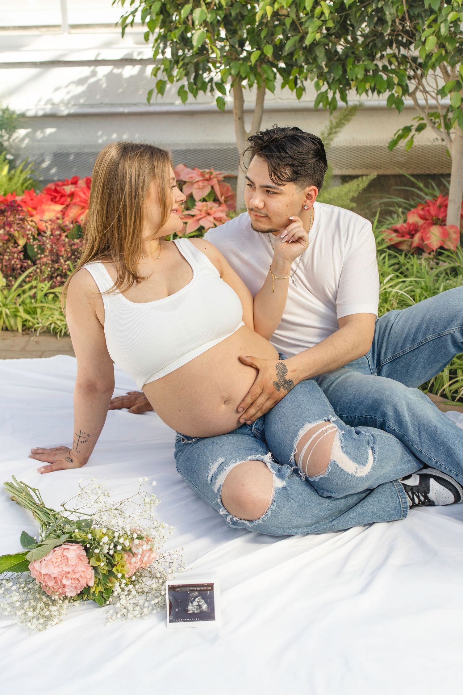 An expectant couple shares intimate moment on white blanket surrounded by flowers in greenhouse.