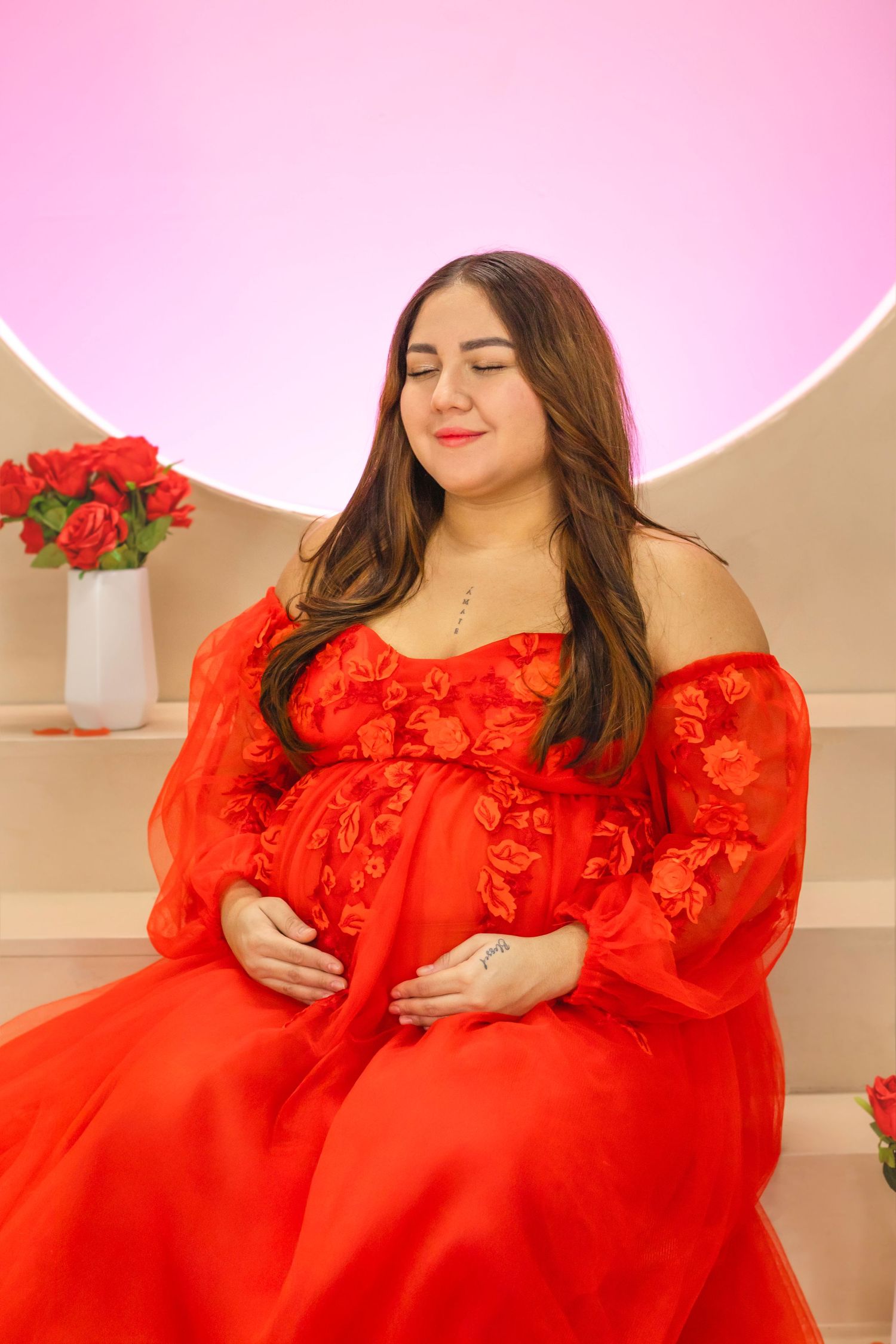 A maternity photoshoot featuring a red lace dress and pink backdrop with roses.