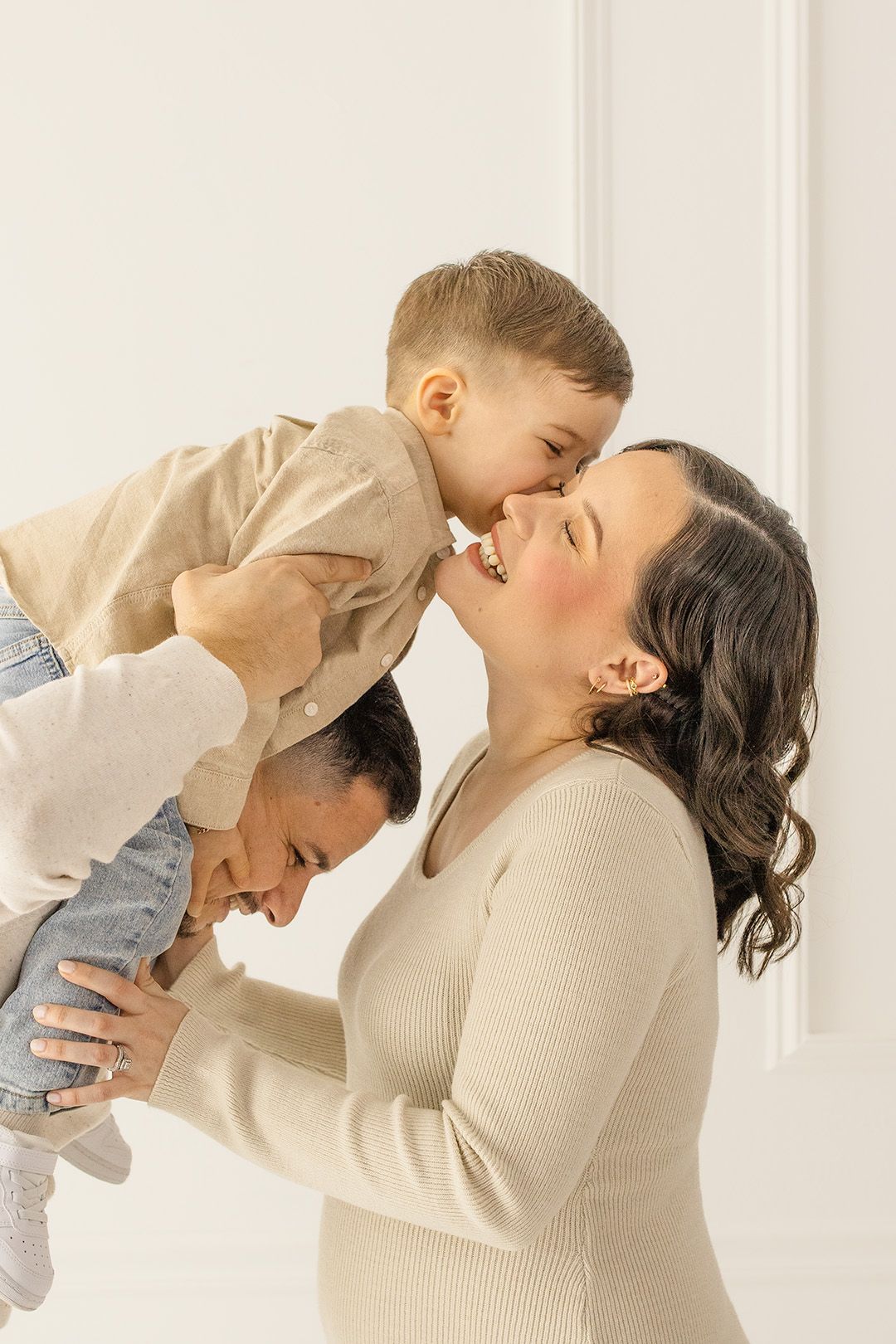 A mother smiles as her young son gives her a kiss while being lifted by his father during a family maternity photo session.