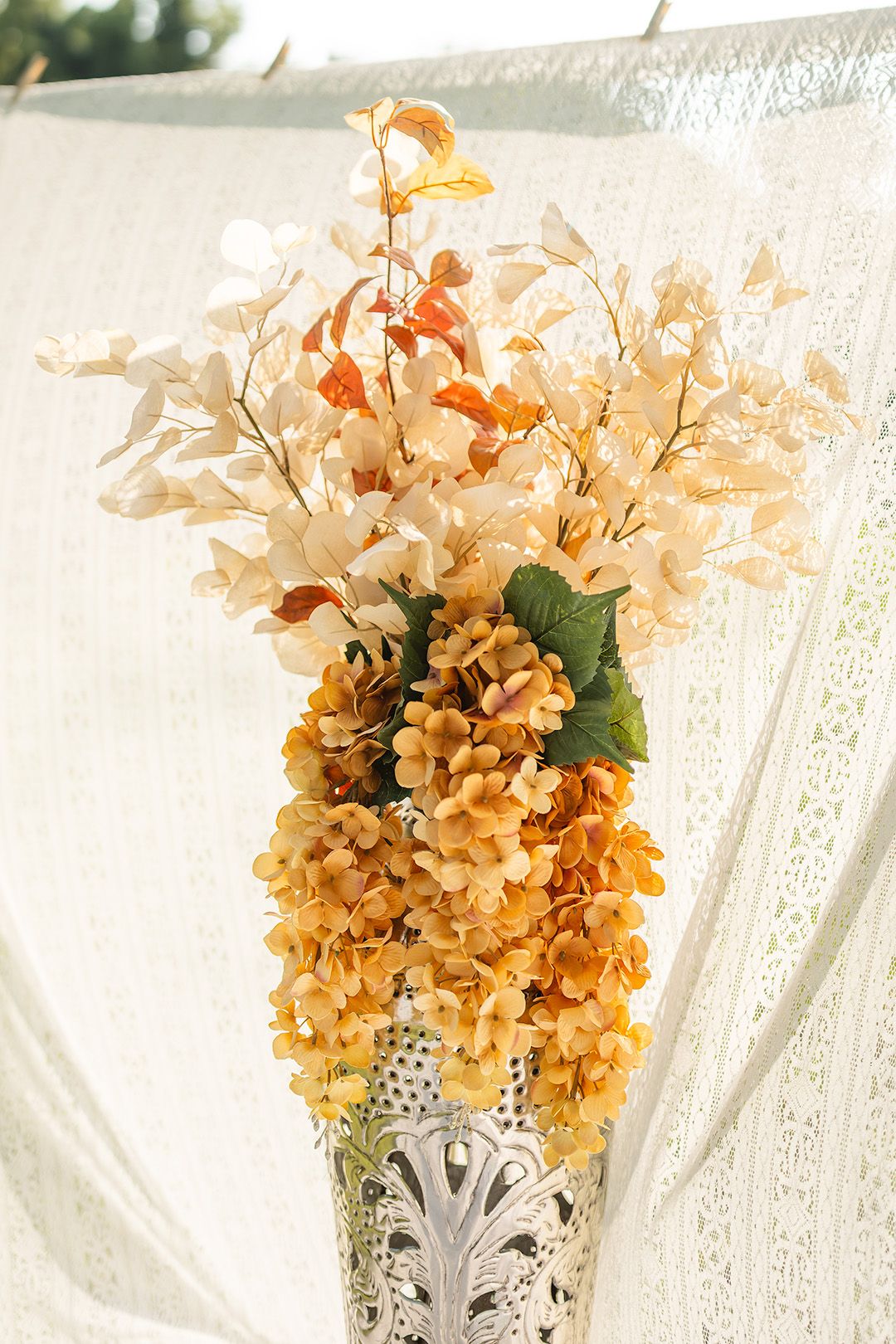 Silver vase with dried hydrangeas and white blooms arranged against a white lace backdrop.