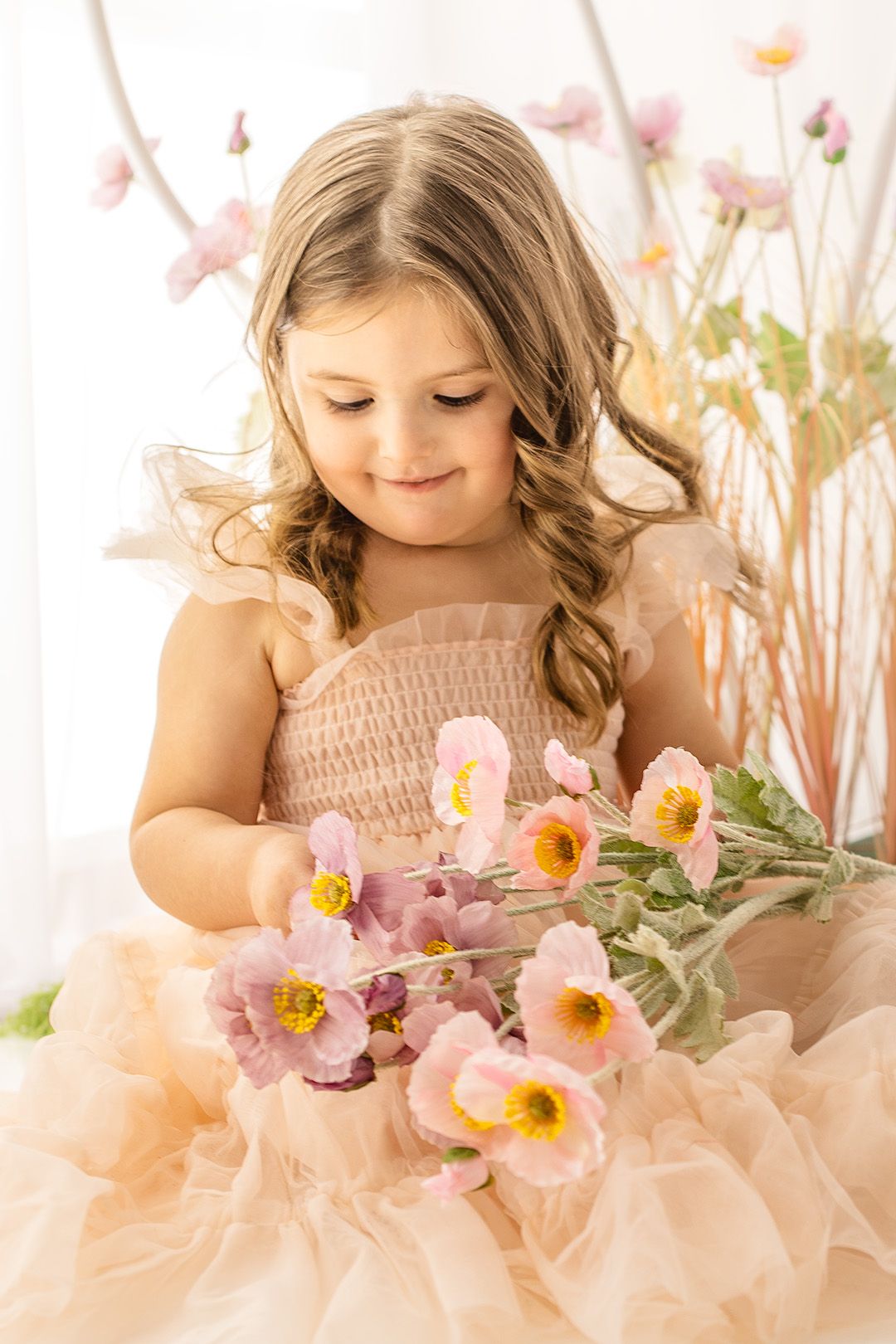A child in a pink tulle dress smiles while holding delicate pink poppy flowers against a light floral backdrop.