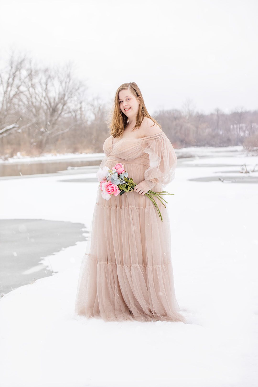 Person wearing blush tulle gown holds pink flowers in snowy winter setting.