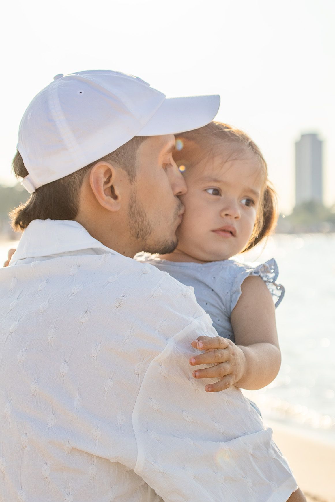 A father tenderly kisses his baby girl on the cheek while holding her at a sunny beach.