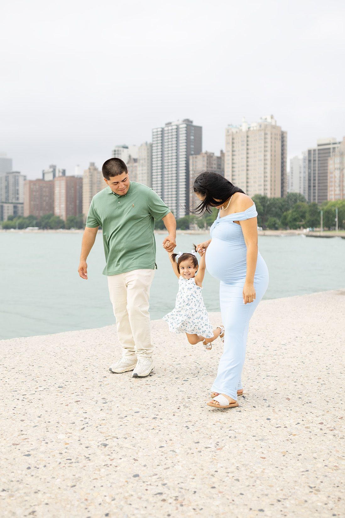 Pregnant woman in blue dress and man in green shirt swing toddler girl between them on a lakeside path with city skyline.