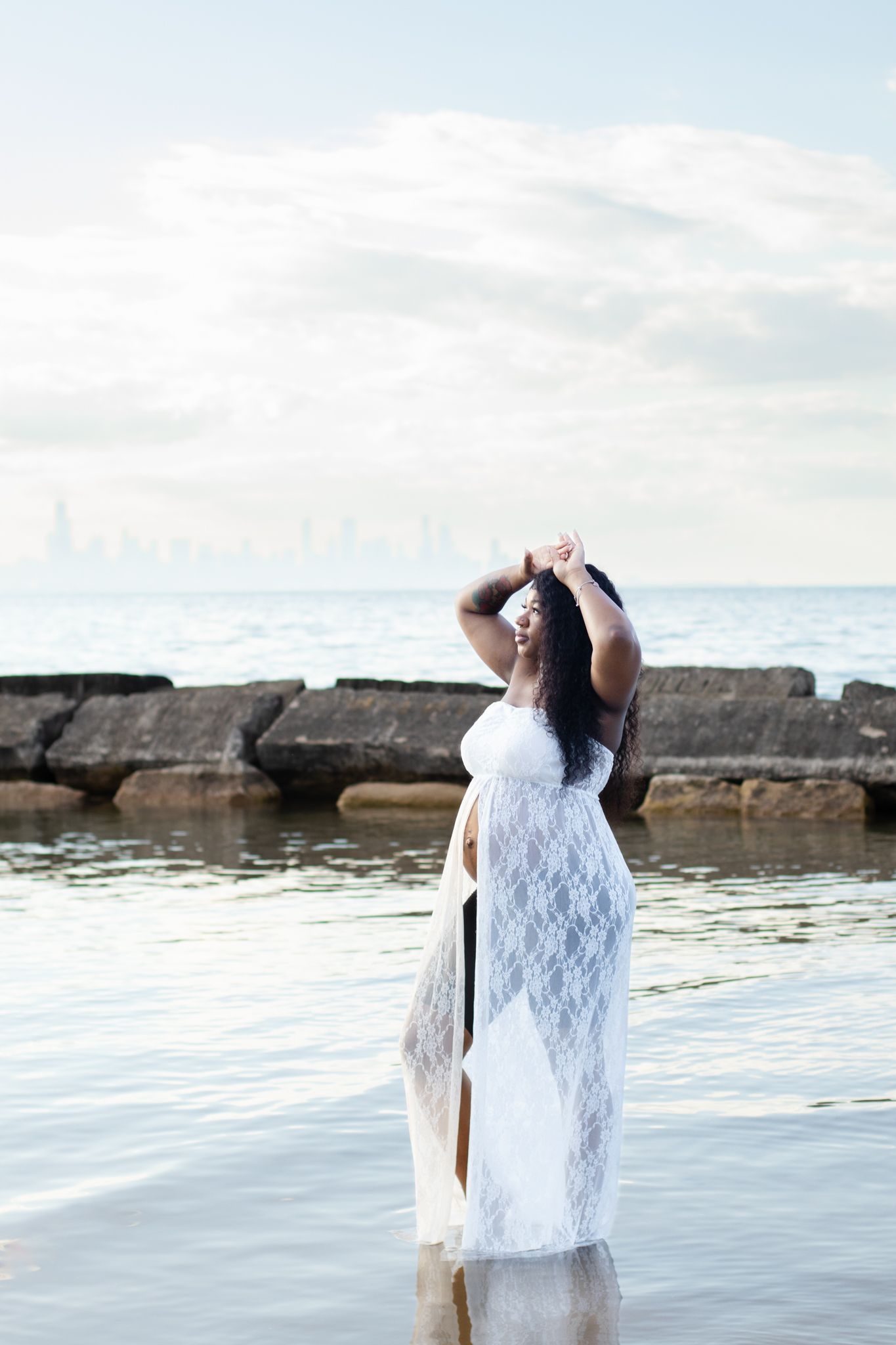 Expecting mother in white lace dress poses in water at beach during maternity photoshoot.