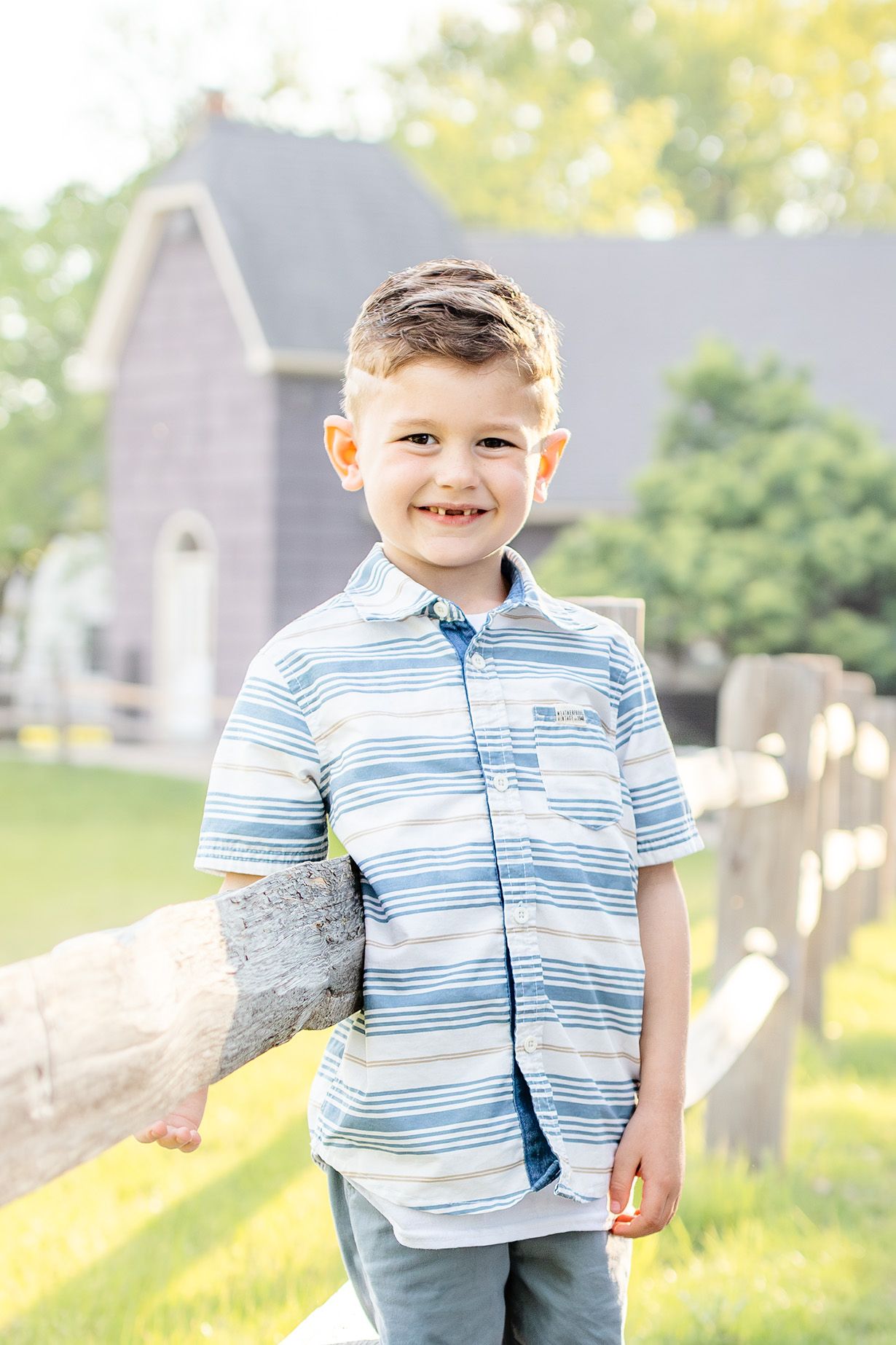 Smiling young boy in striped blue shirt standing by wooden fence with green yard and house in background.