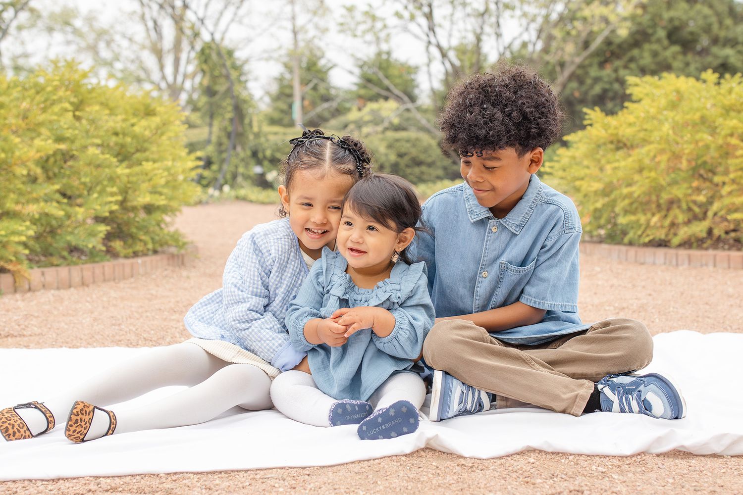 Three children in blue outfits sit together on a white blanket outdoors, smiling warmly in a park setting.