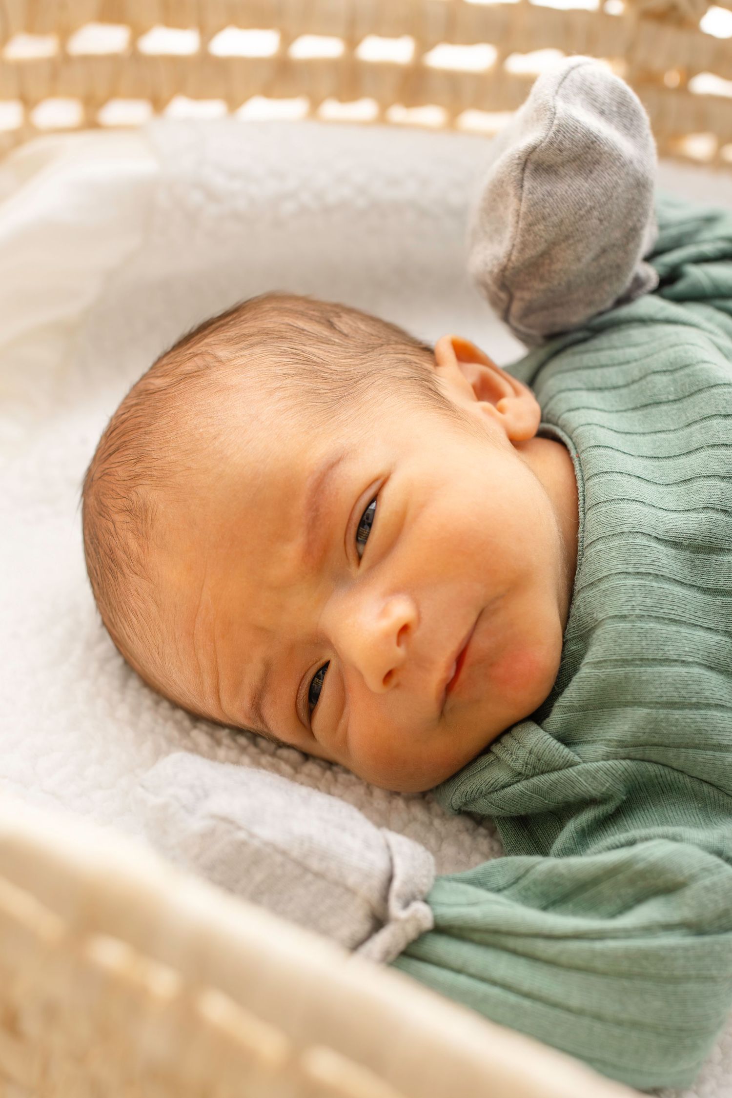 Baby resting on white bedding while wearing a sage green sweater and gray hat.