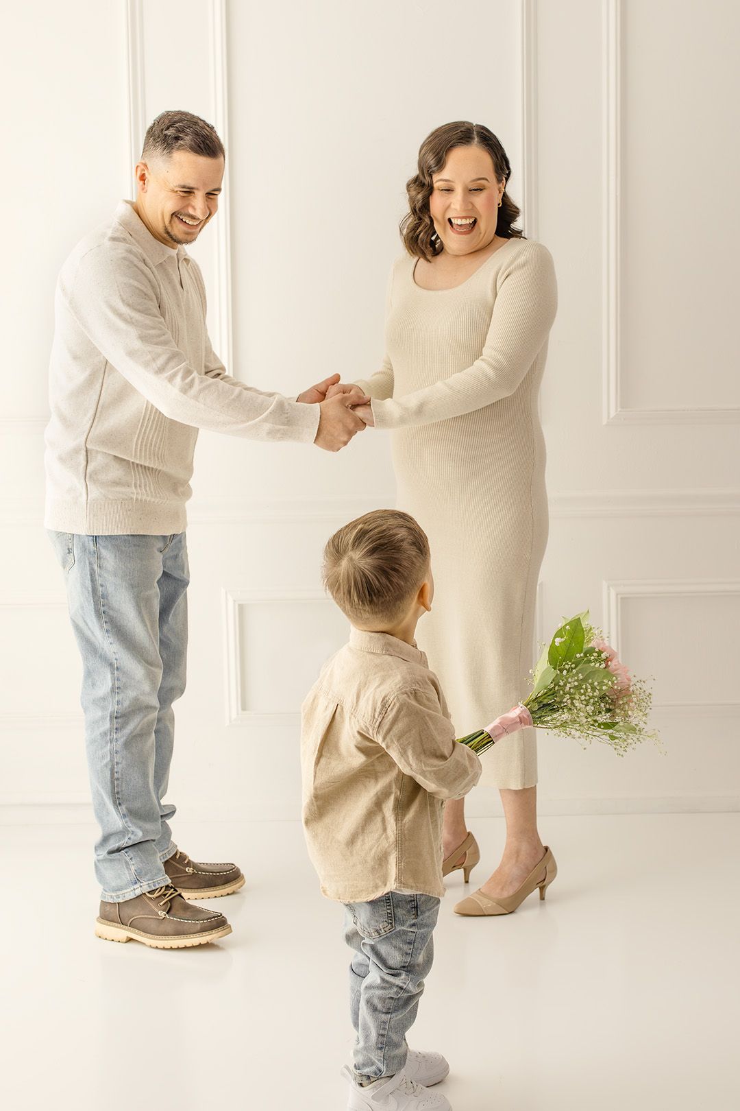 Happy pregnant woman receives flowers from toddler son while husband holds her hands in bright white room.
