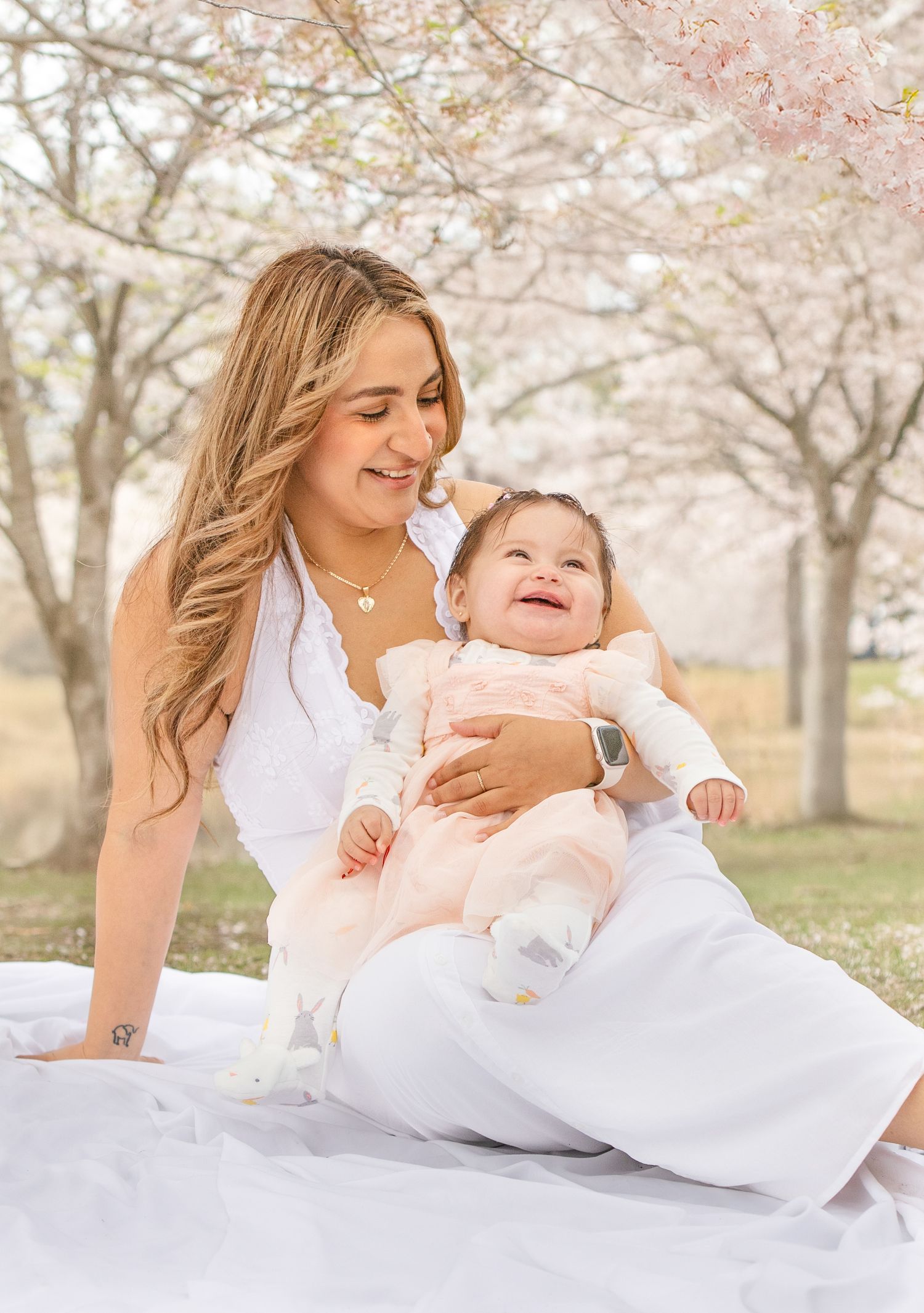 A tender moment shared between parent and baby under blooming cherry blossom trees on a white blanket in springtime.