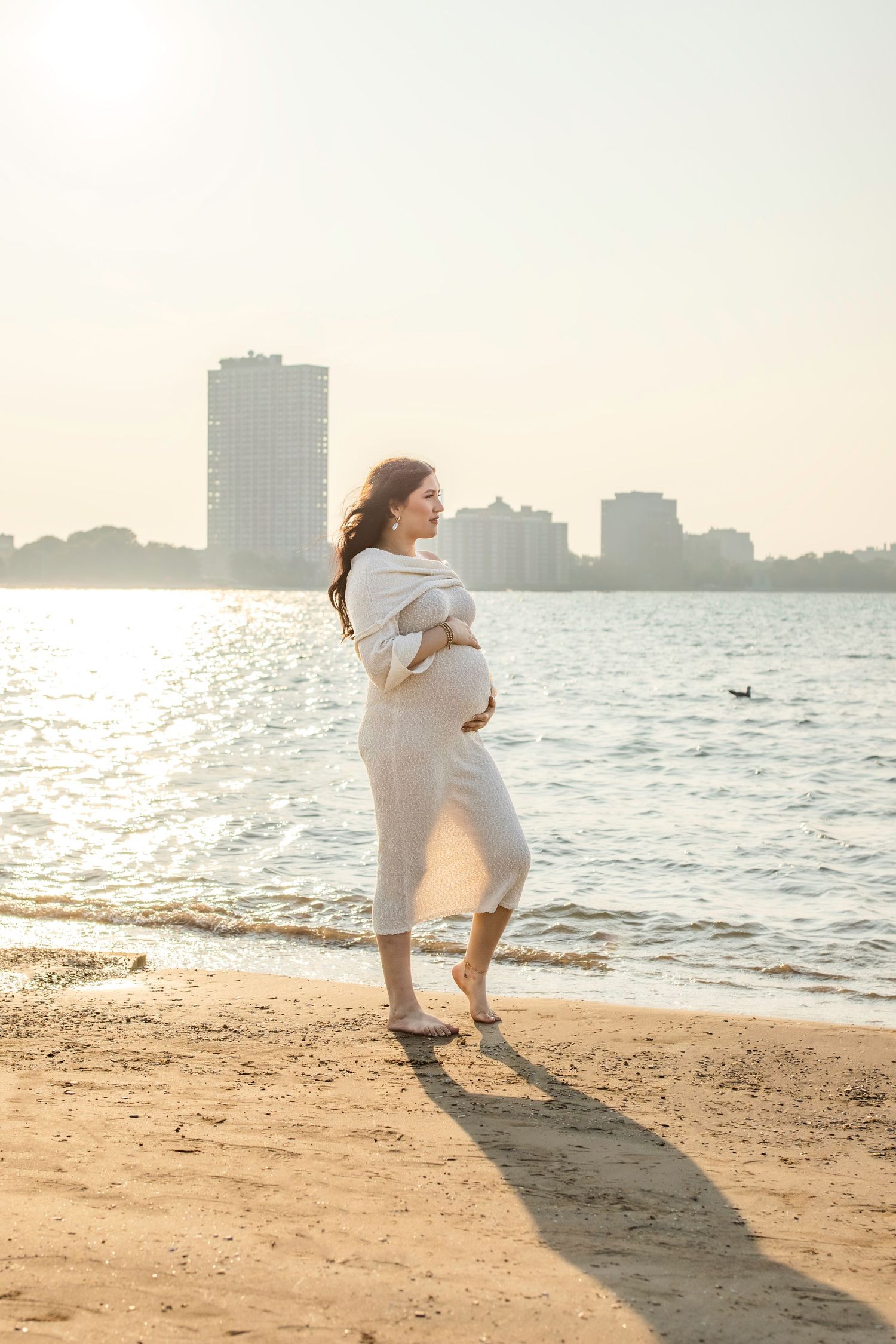 Pregnant woman in white dress standing on sunny beach holding belly with city skyline and water in background.