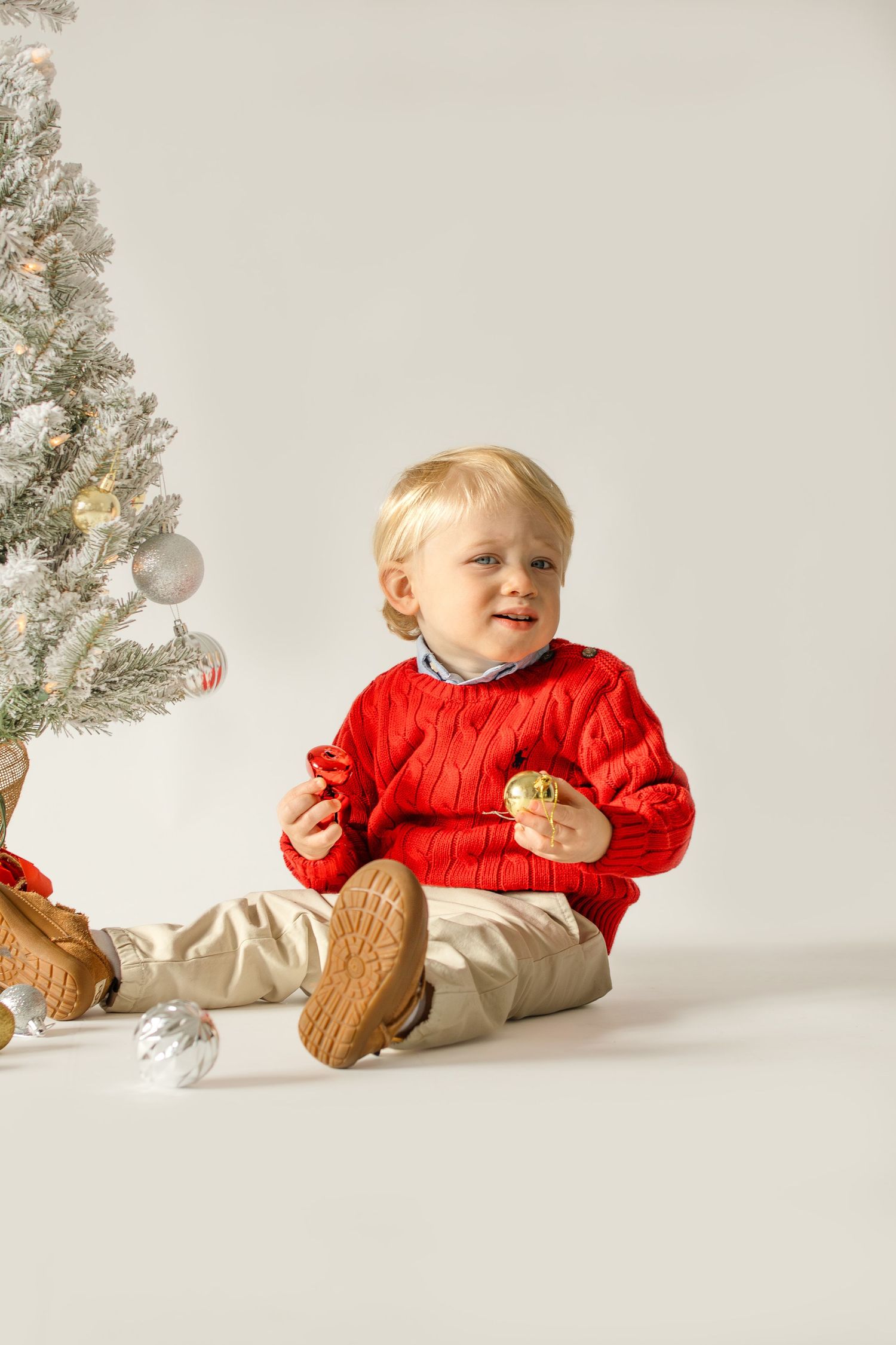 A small child in a festive red sweater crawls and plays near a silver Christmas tree decoration.