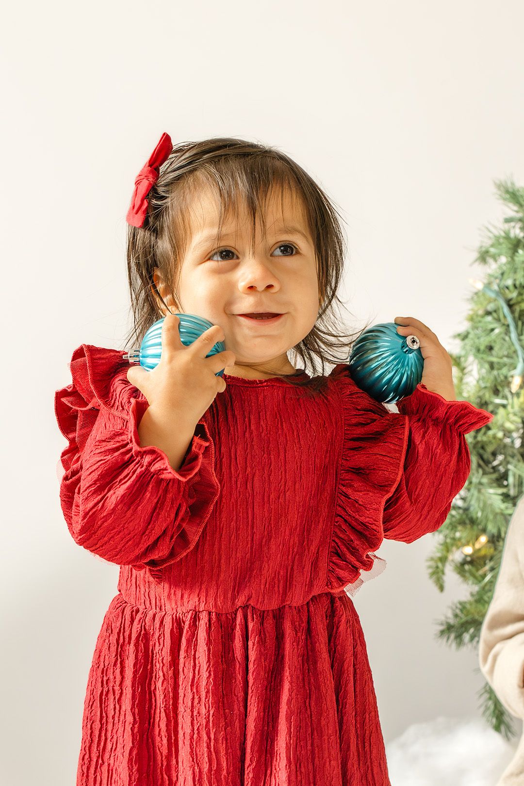 Child in red holiday dress holds blue ornaments while playing and posing against white background.