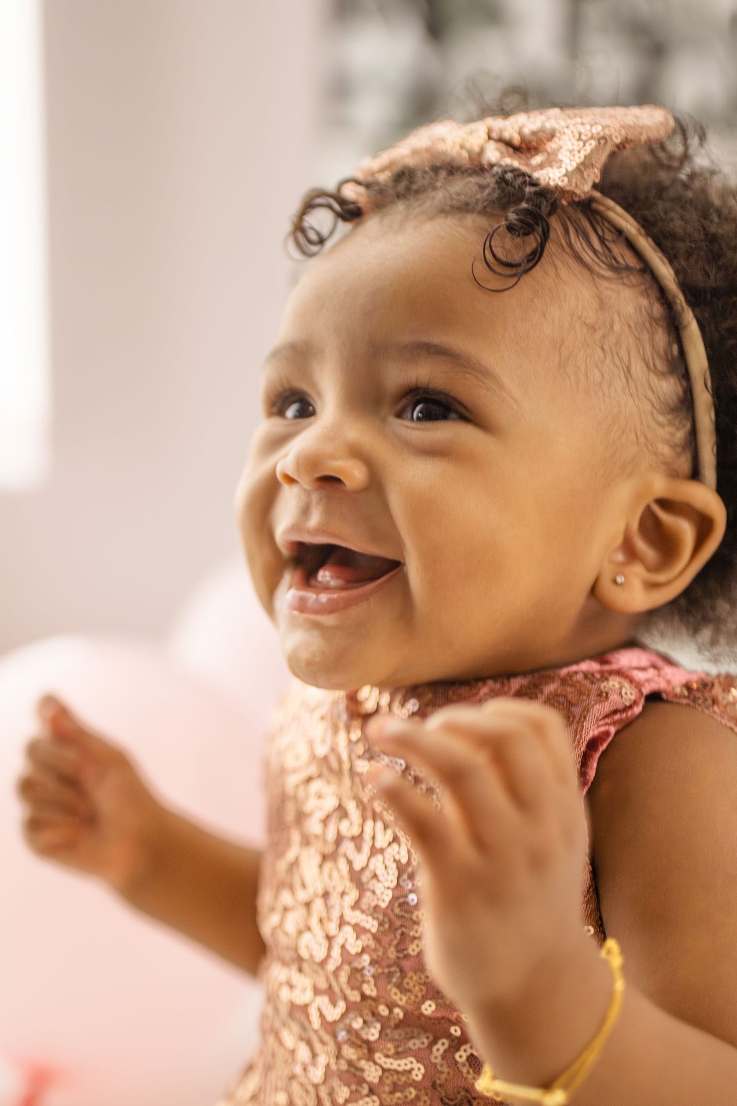 Joyful baby girl in rose gold sequin dress and bow headband laughing with tiny fists raised.