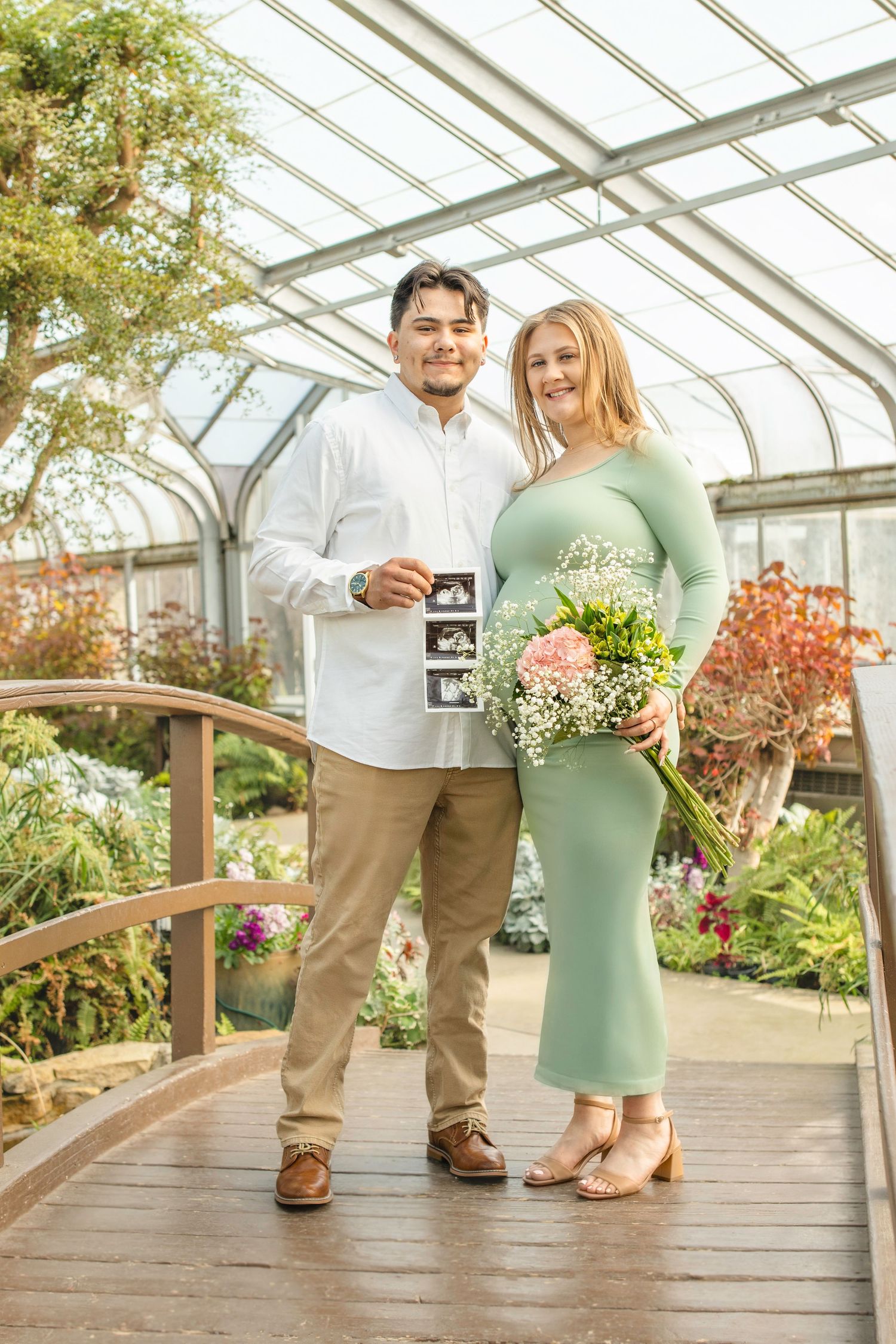 Expecting couple holding ultrasound photo and flowers in greenhouse setting with natural lighting.
