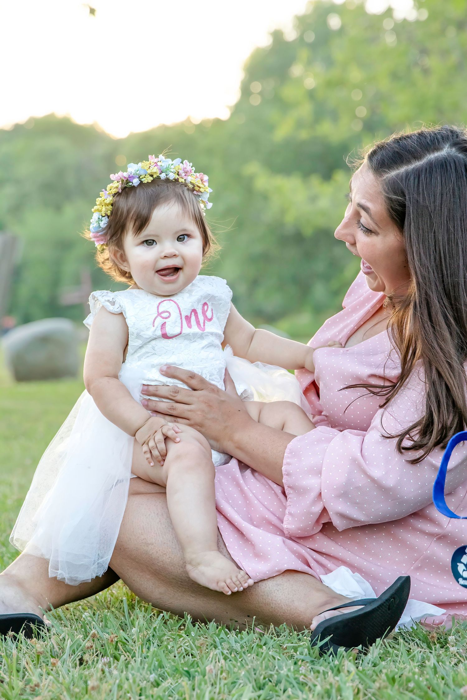 Mother holds smiling baby girl wearing floral crown and “One” outfit at first birthday party.