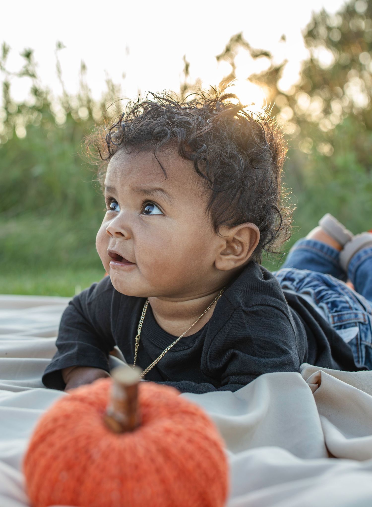 A small child on a white blanket looking up at golden sunlight next to an orange pumpkin during autumn.