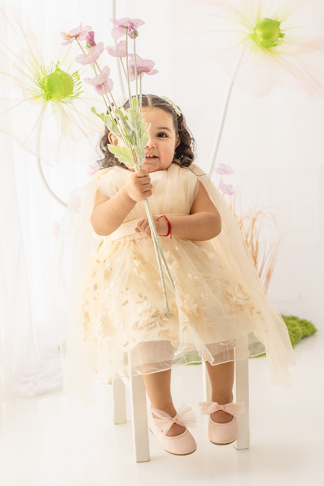 A toddler girl in a cream tulle dress holds pink flowers while sitting on a white stool in a floral studio setting.