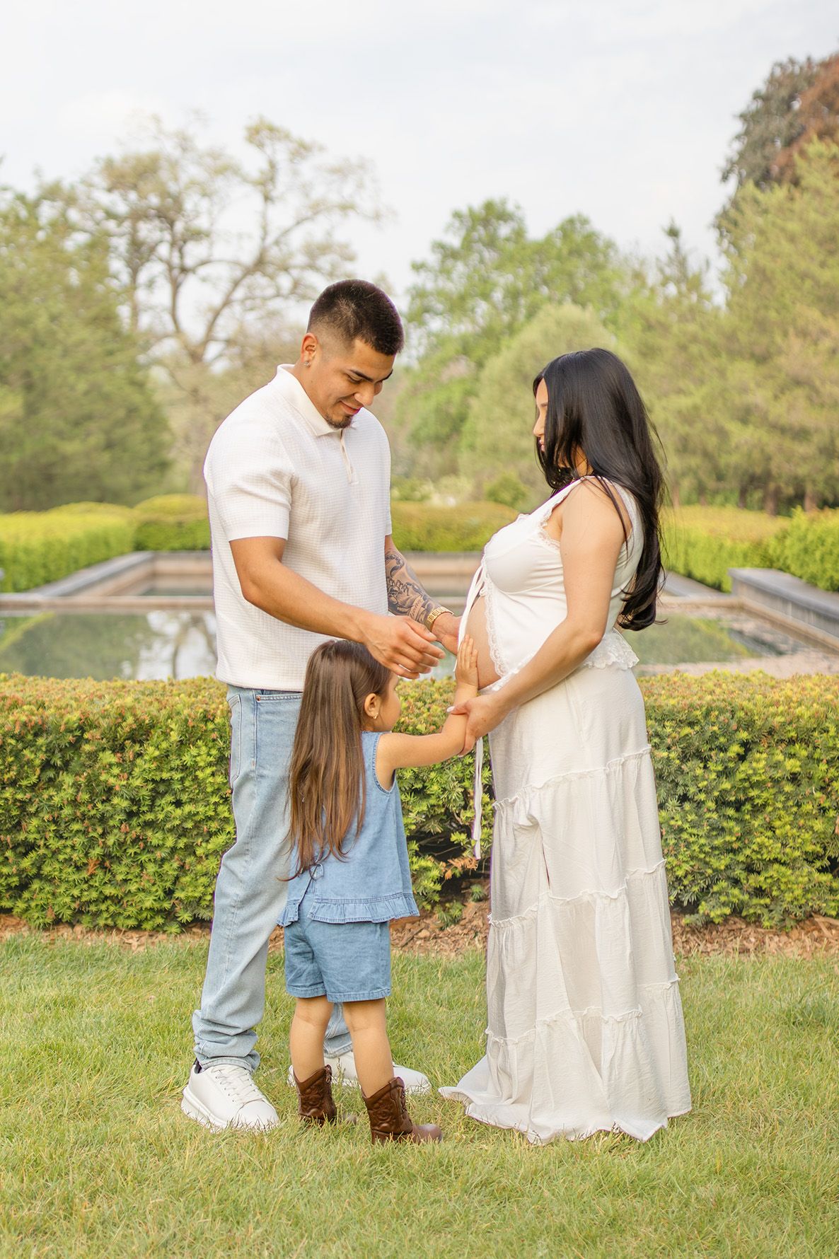 A family maternity photo with a man, pregnant woman in white dress, and young girl in a lush green garden.