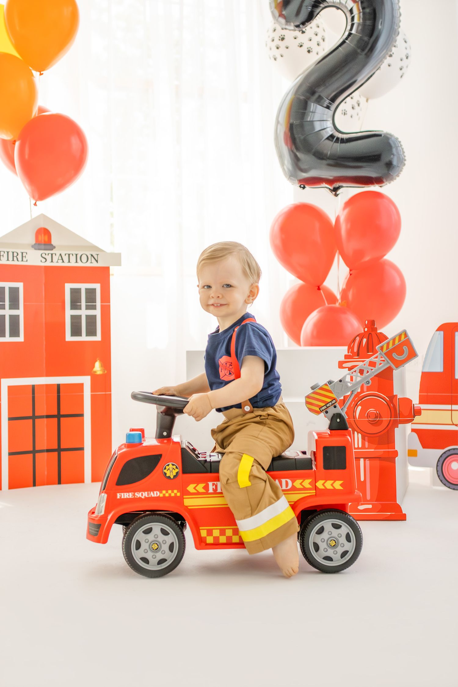 Toddler boy riding red toy fire truck at 2nd birthday party with balloons and fire station decorations.