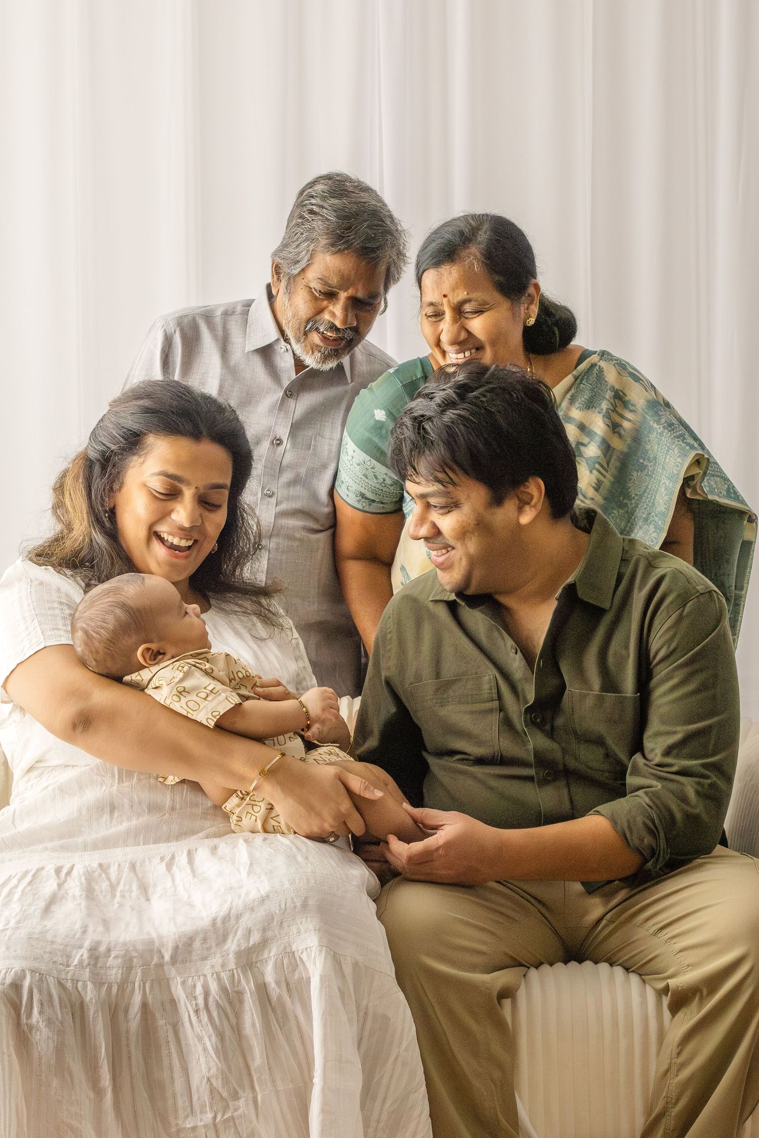 Multi-generational Indian family shares joyful moment with baby in white room setting.
