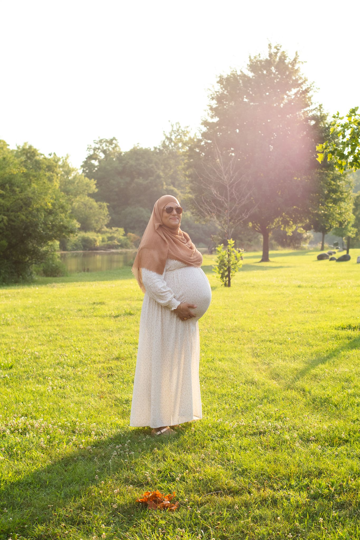Expectant mother in flowing white dress poses in sunlit garden setting.