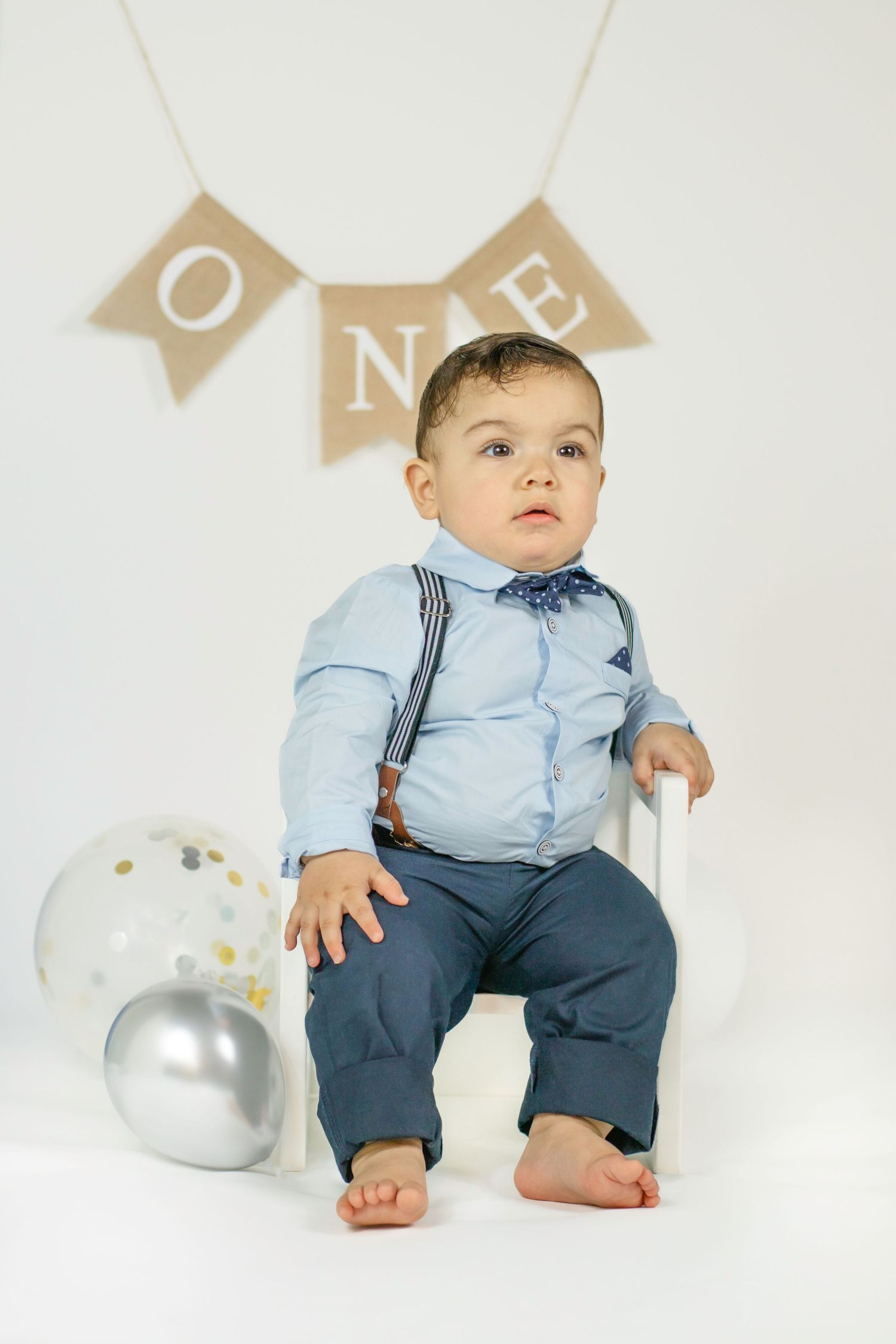 A baby boy in suspenders and bow tie sits for his first birthday photo shoot with balloons and ONE banner.