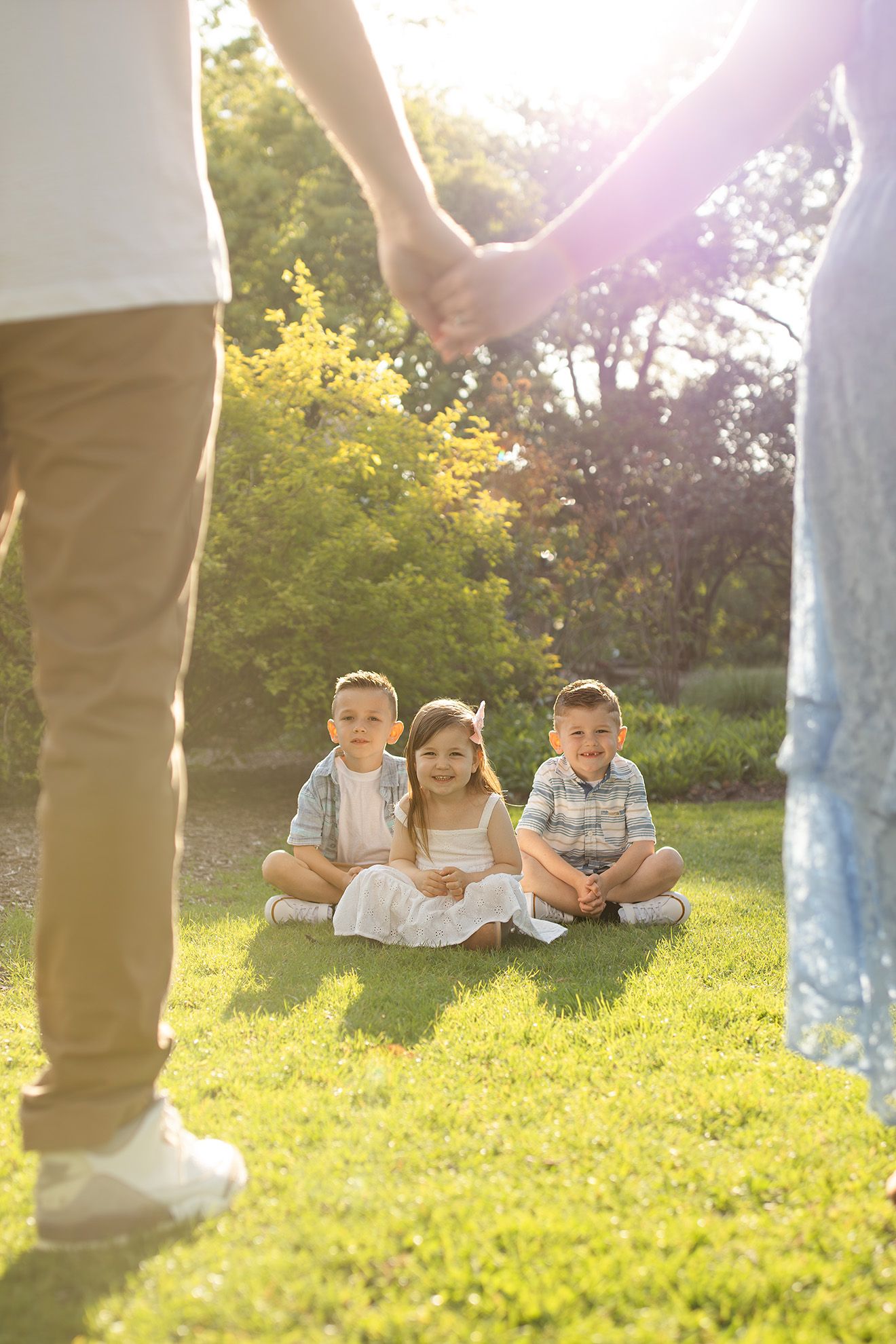 Three children sitting on grass in a sunny park while their parents hold hands above them.