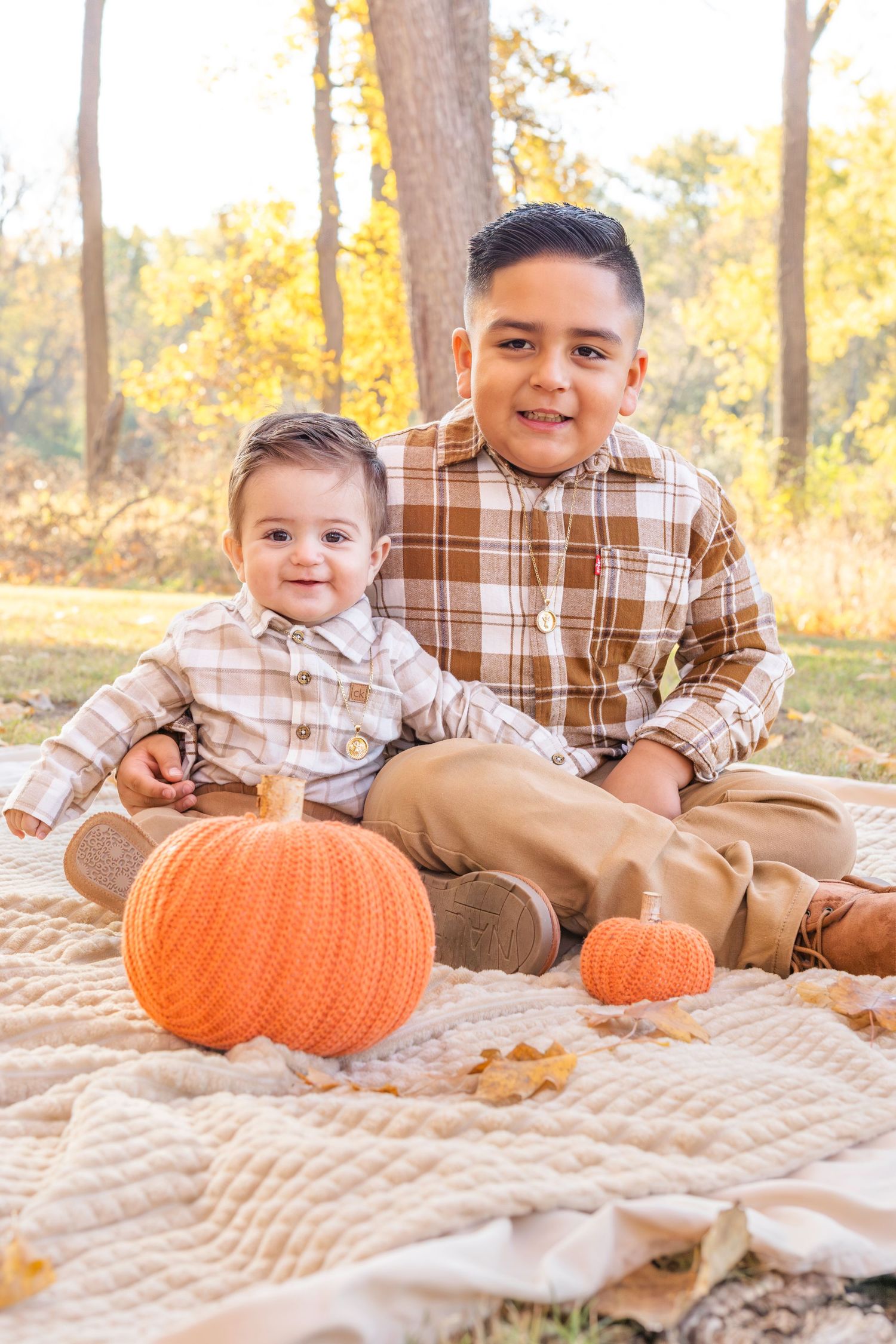 Three children sit on a blanket with pumpkins during an autumn outdoor photo session in matching plaid outfits.
