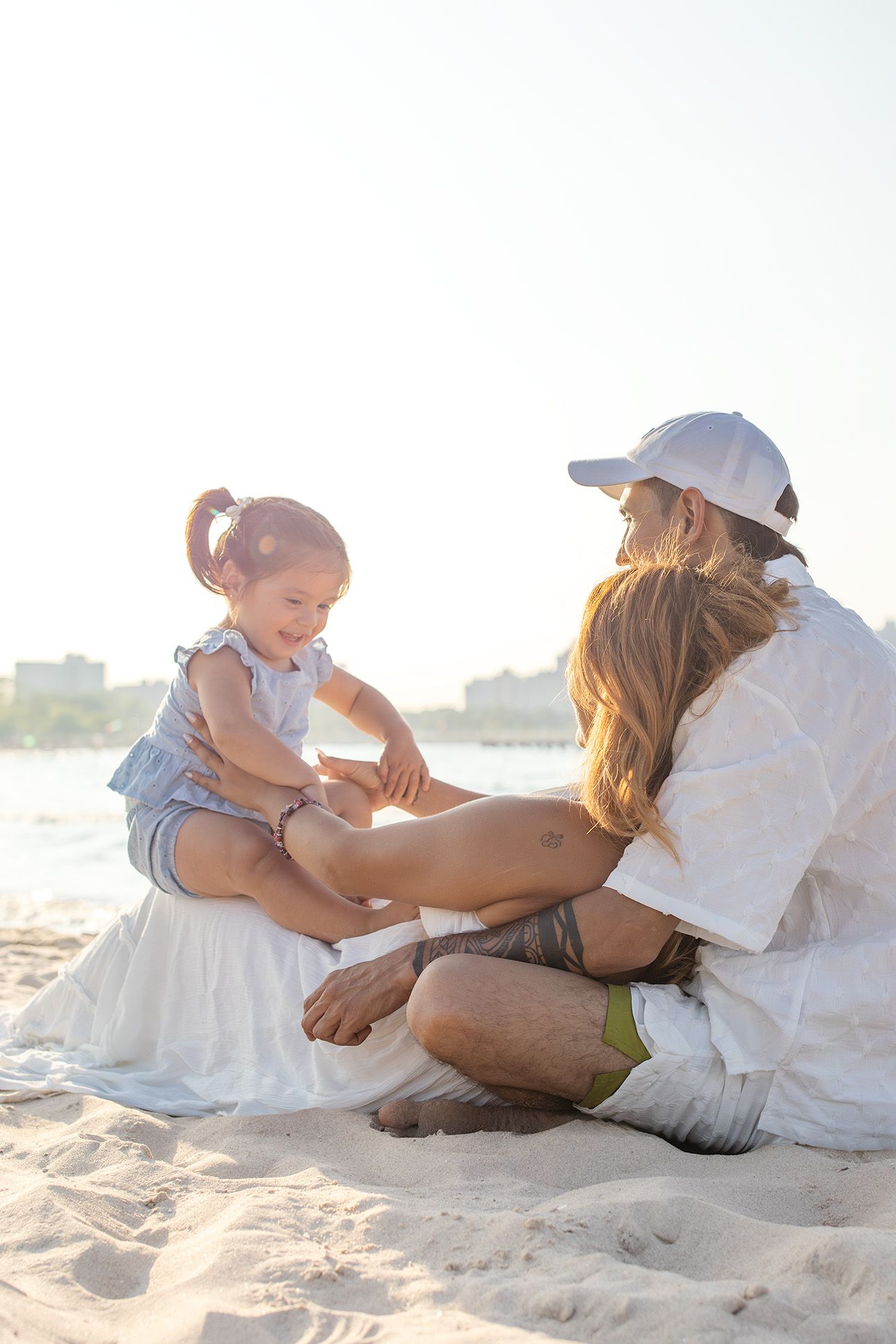 Young family sitting together on sunny beach with toddler girl in blue dress smiling.