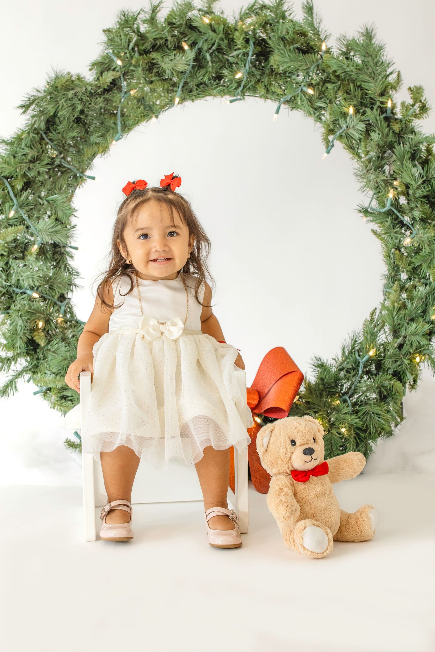 A festive holiday scene with a white dress, teddy bear, and evergreen Christmas wreath against a white background.