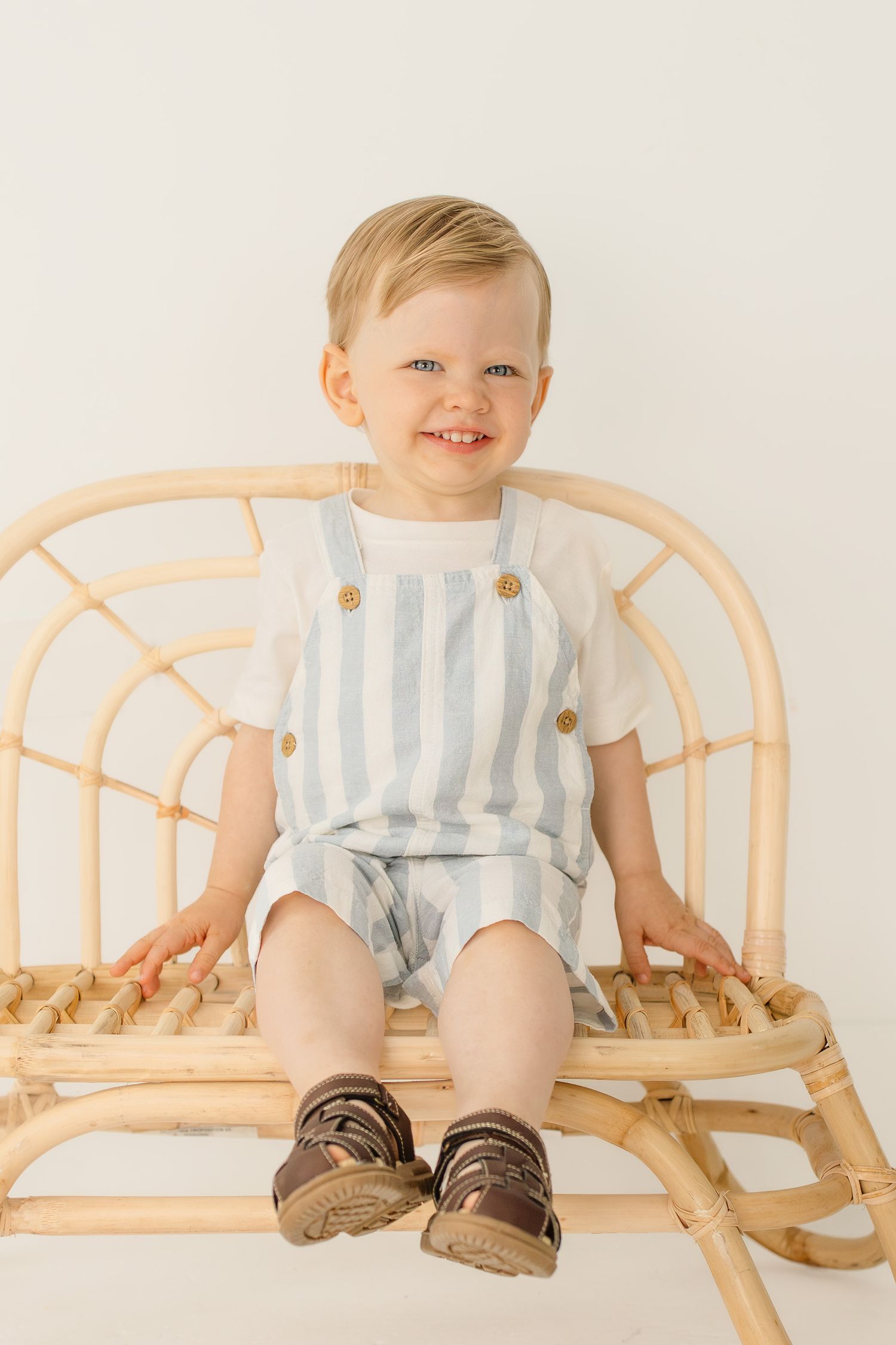 Happy toddler boy wearing striped overalls and brown sandals sits smiling on a rattan bench.