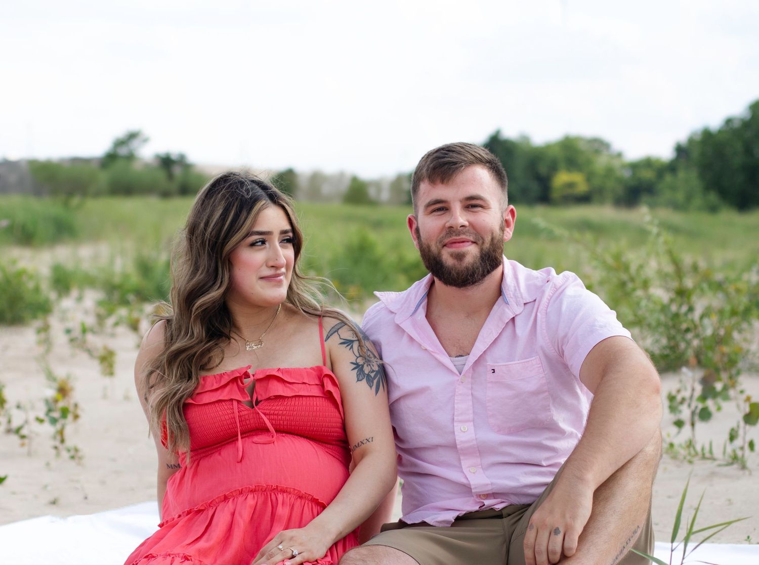 Couple sitting together on beach wearing pink shirt and red dress during casual photoshoot.