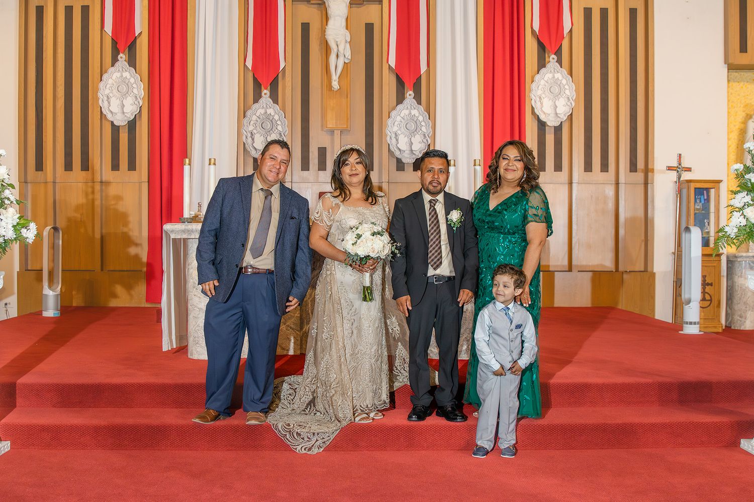 Wedding party posing in church with red carpet, bride in lace gown, groom, family members in blue suit and green dress.