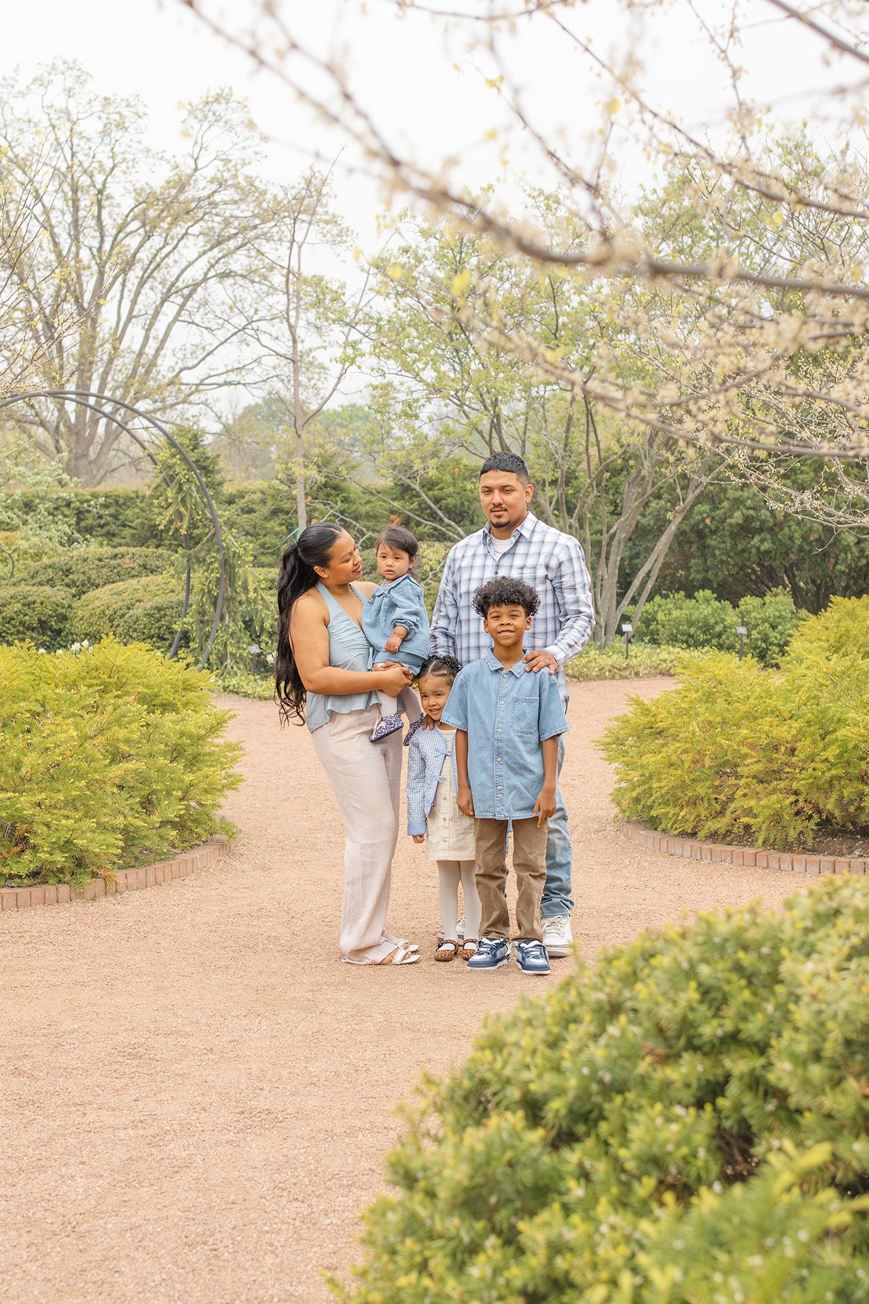Family of five poses on a garden path surrounded by lush greenery, dressed in coordinating blue and neutral outfits.