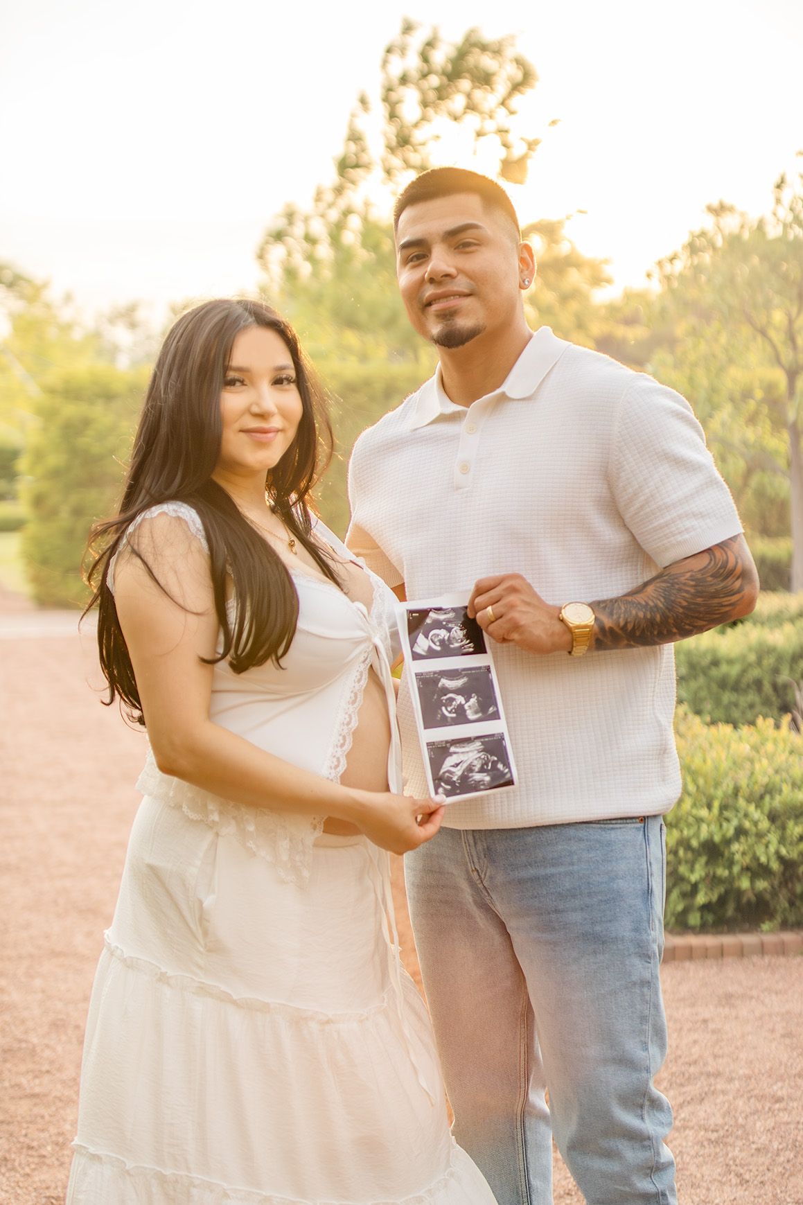Expecting couple in white outfits holds ultrasound photos together during a sunny outdoor maternity photo session.