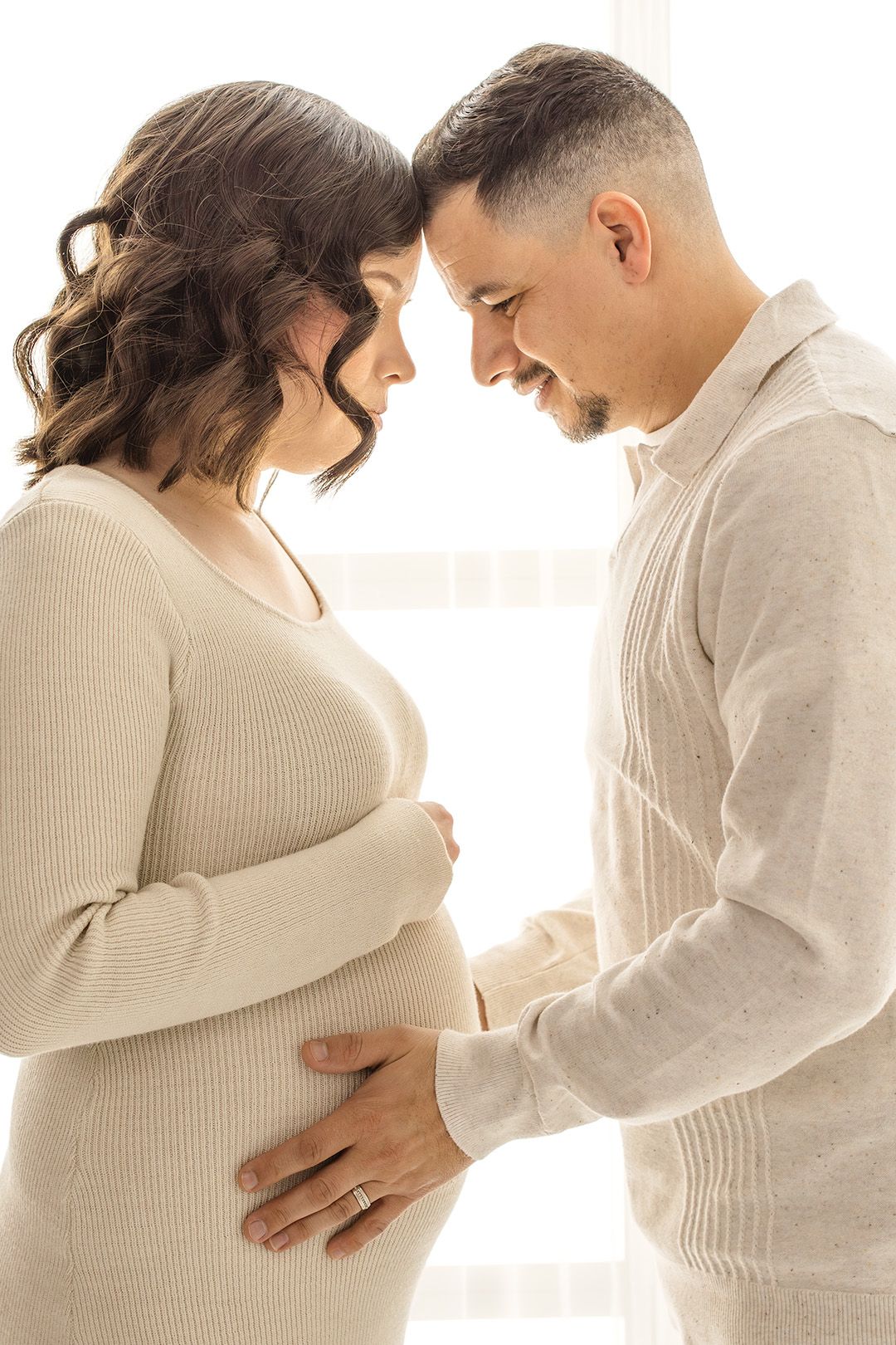 A pregnant woman and her husband tenderly touch her baby bump while standing forehead to forehead in a bright studio.