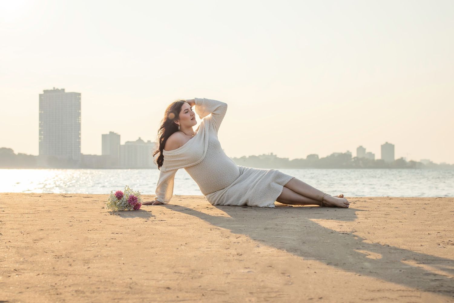 Two people in white outfits pose intimately together on a sandy beach at sunset with city skyline in background.