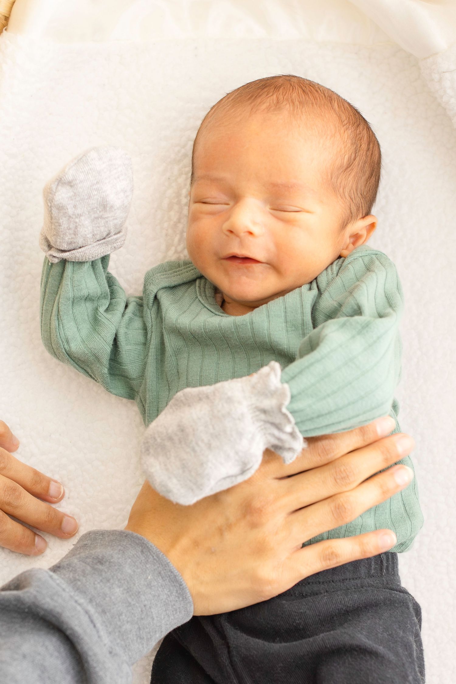 A peaceful sleeping newborn wearing a mint green sweater and gray pants lies on a white blanket.