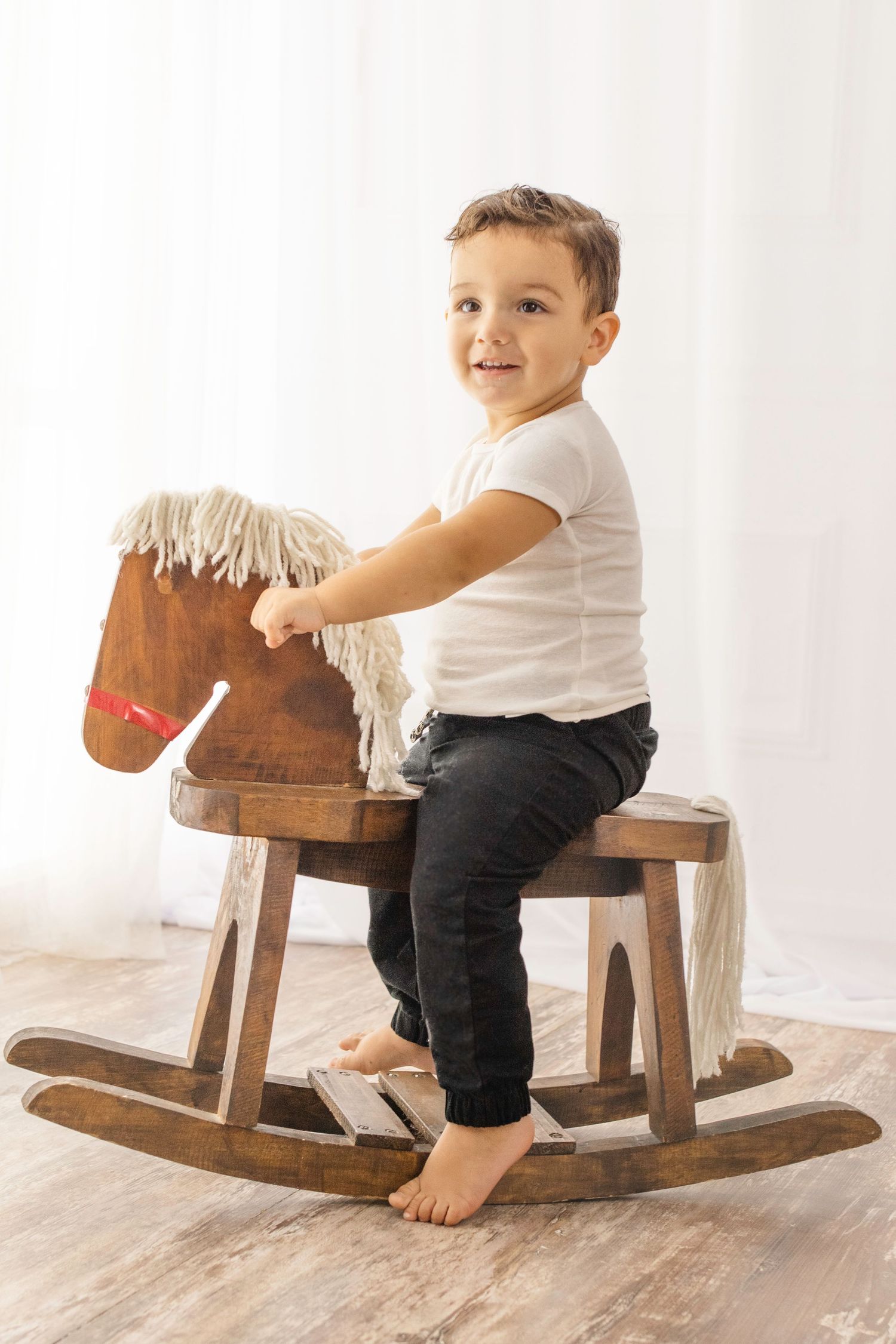 A child plays on a wooden rocking horse toy in a bright white room with hardwood floors.