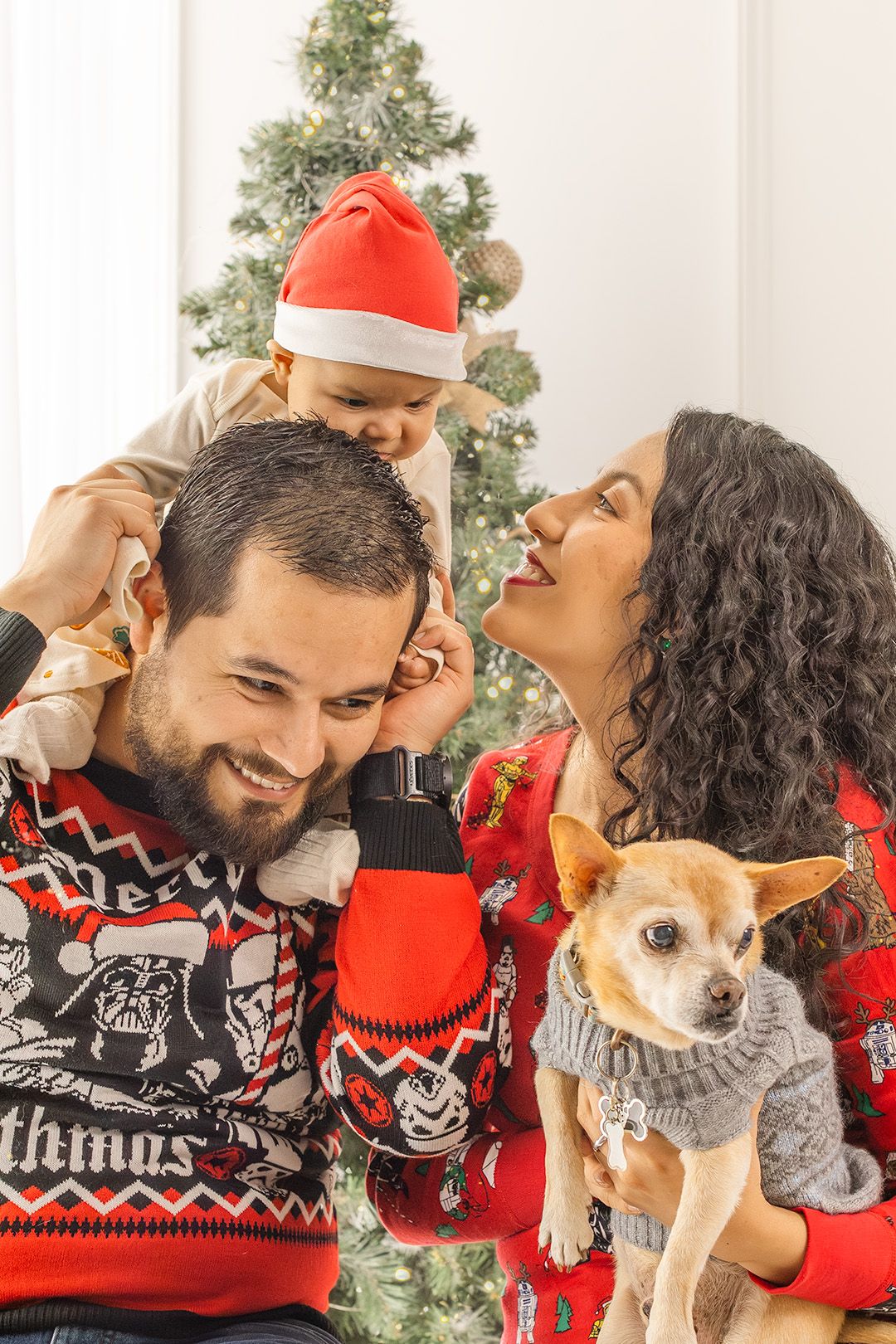 Family in Christmas sweaters and Santa hats cuddling with their small dog during holiday photoshoot.