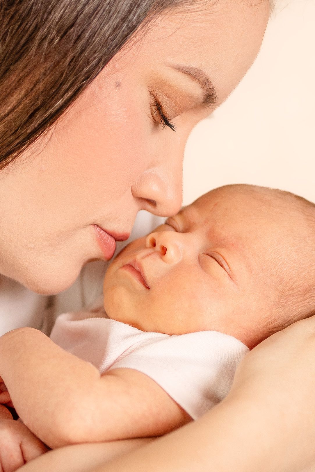 A tender mother gently kissing her sleeping newborn baby, captured in a warm, intimate close-up portrait.