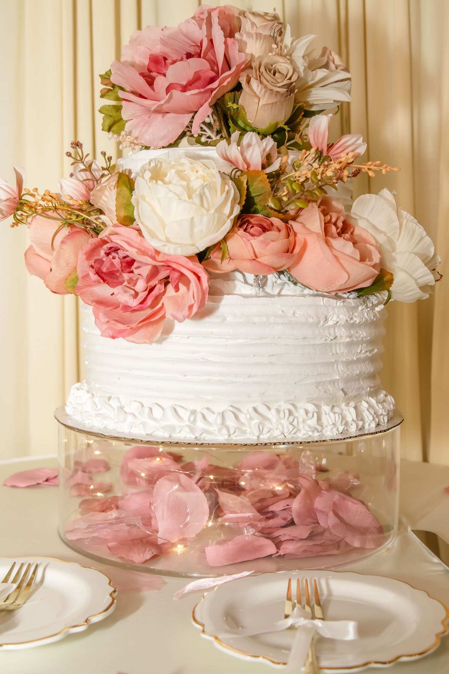 An elegant white wedding cake decorated with pink and peach roses and petals displayed on a crystal cake stand.