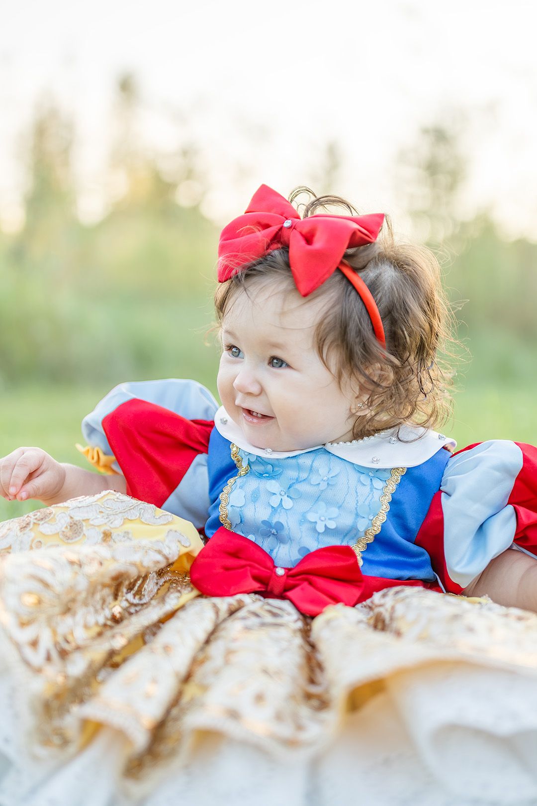 Baby girl in Snow White costume with red bow headband sitting outdoors in green field.