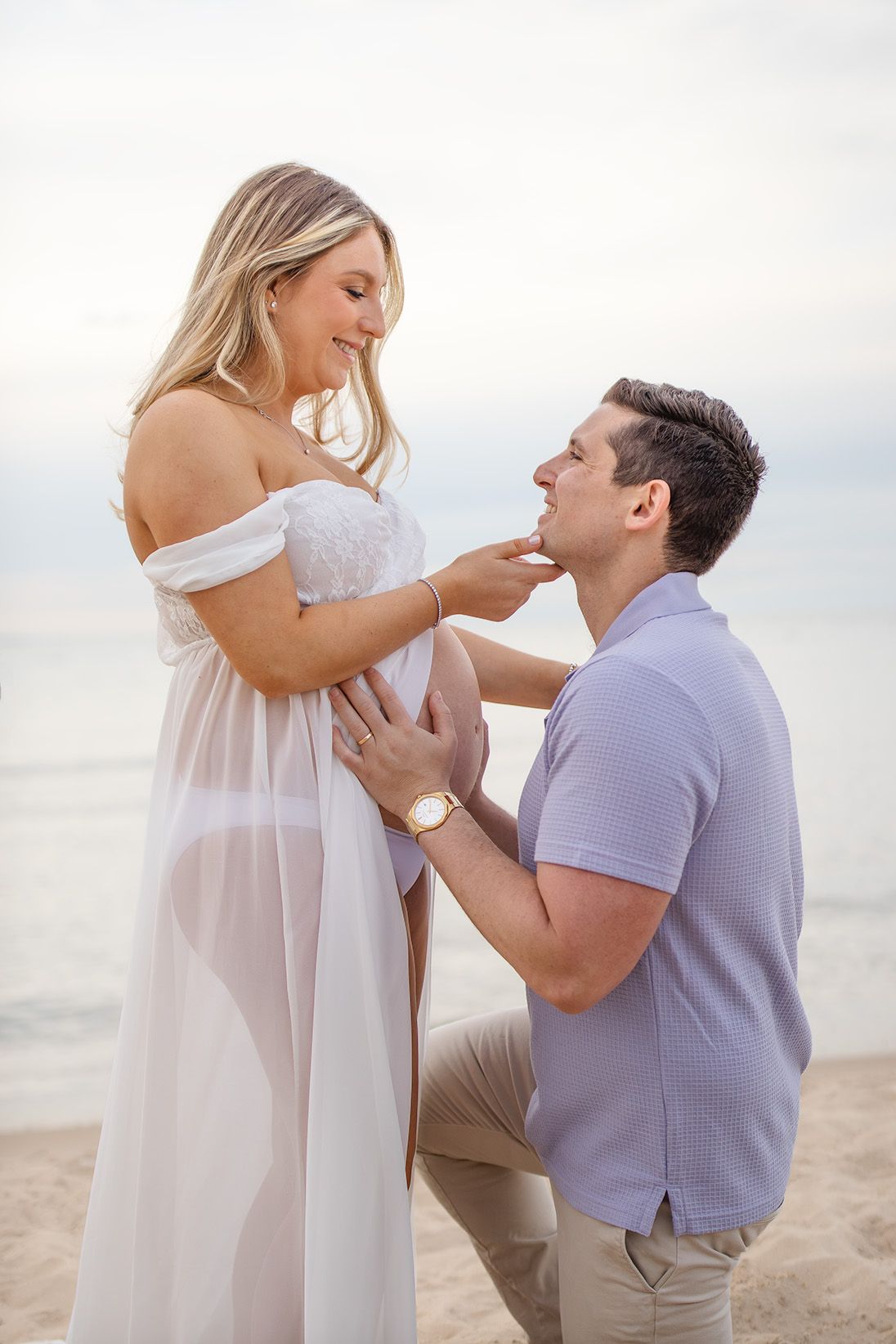 Pregnant woman in white dress with man kneeling on beach, touching her face in a tender maternity photo shoot.